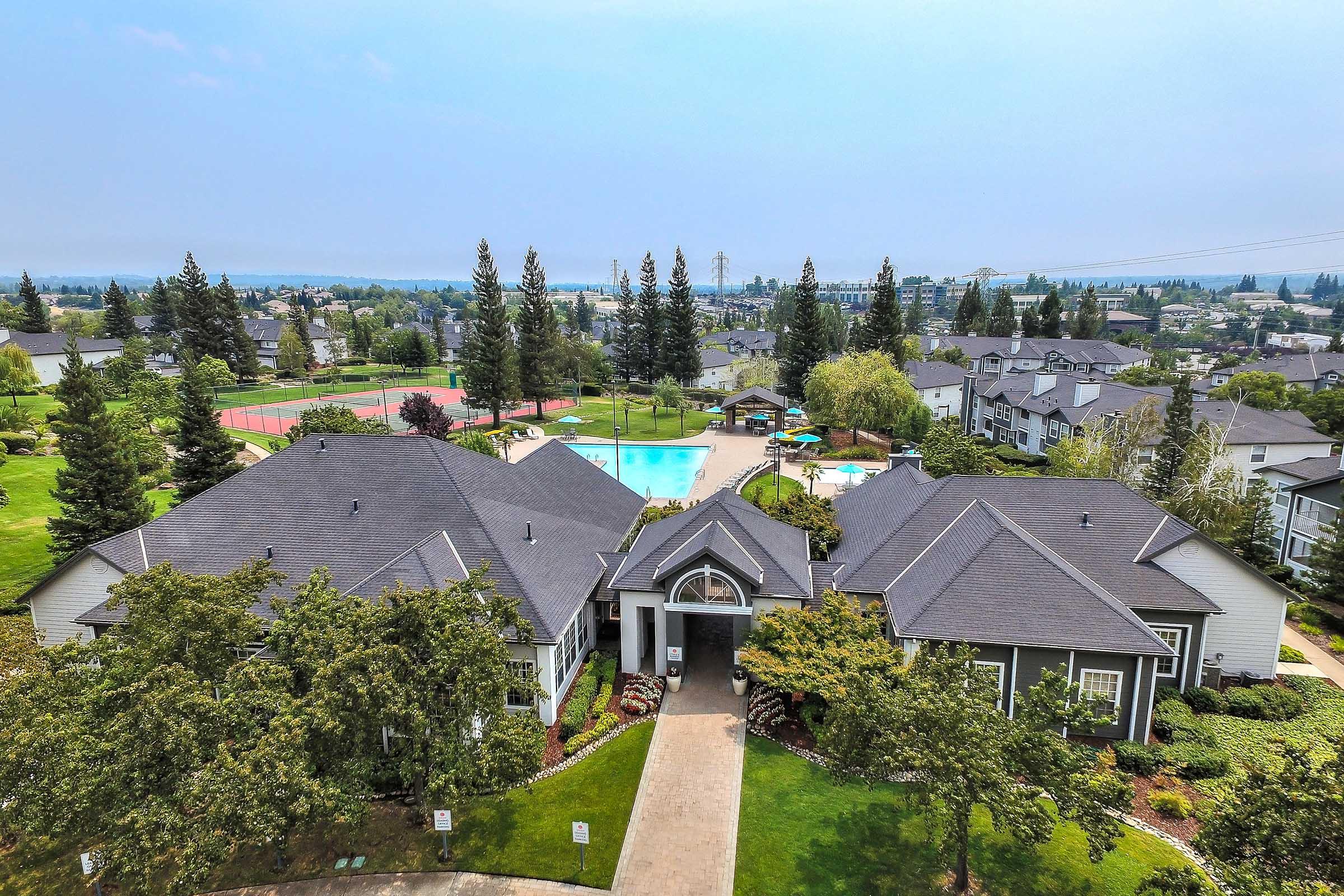 Aerial view of a residential complex featuring a central clubhouse, landscaped greenery, and a swimming pool surrounded by trees. Nearby amenities include tennis courts and walking paths, with a scenic backdrop of rolling hills and distant power lines.