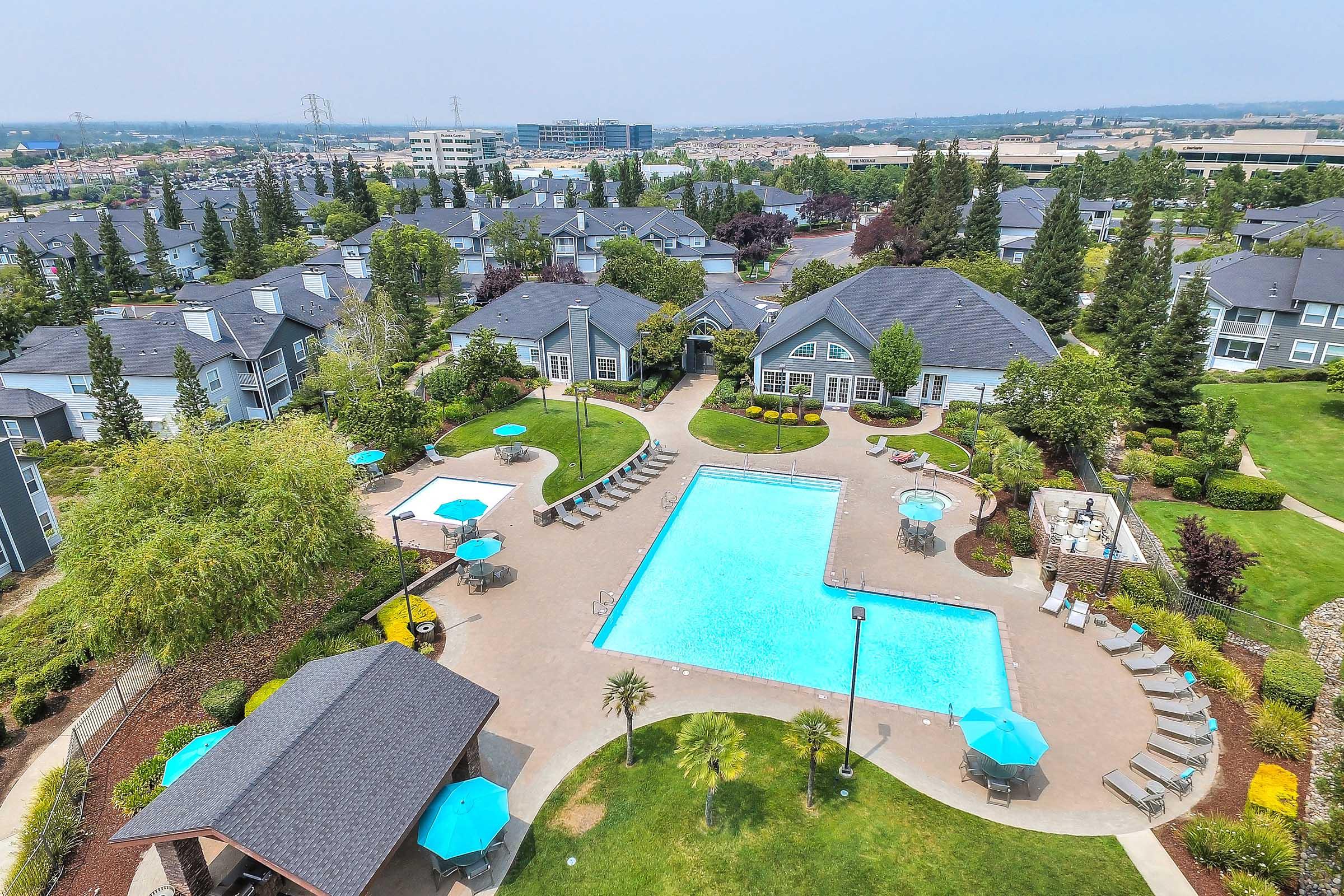 Aerial view of an apartment complex featuring a large swimming pool surrounded by lounge chairs and umbrellas. Lush green landscaping and multiple buildings with gray roofs are visible in the background, along with trees and pathways, creating a serene outdoor environment.