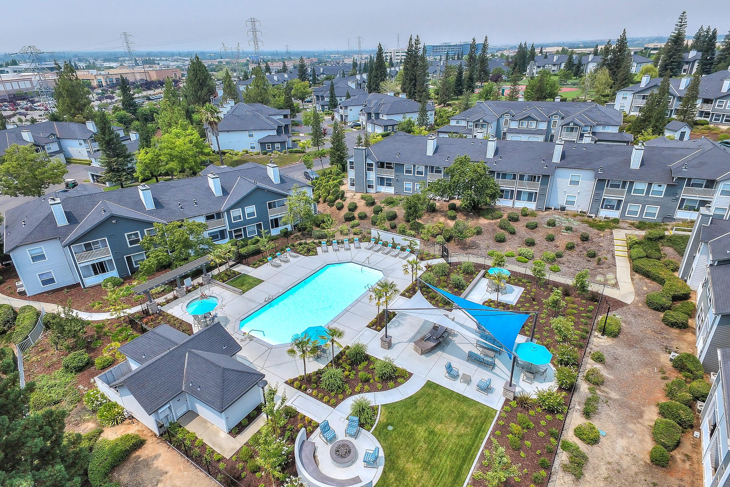 Aerial view of an apartment complex featuring a large swimming pool surrounded by landscaped gardens and lounging areas. Several buildings are visible, set against a backdrop of trees and power lines in the distance. The scene is bright and well-maintained, showcasing outdoor amenities.