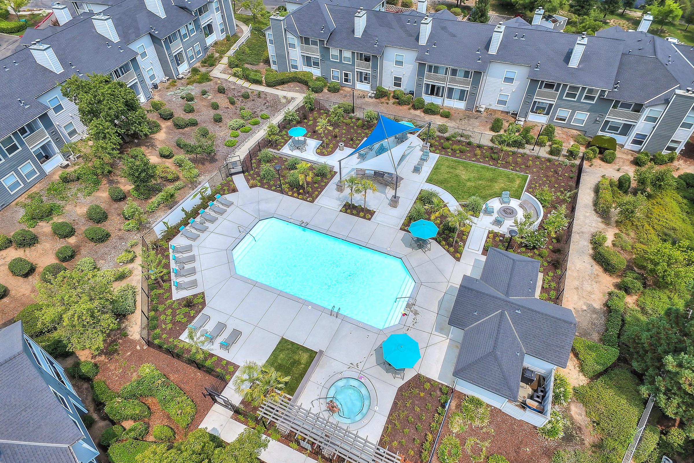 Aerial view of a residential complex featuring a large swimming pool surrounded by lounge chairs and umbrellas. Lush landscaping with shrubs and walkways is visible, alongside a hot tub and outdoor seating areas. Modern apartment buildings frame the pool area, emphasizing a community atmosphere.