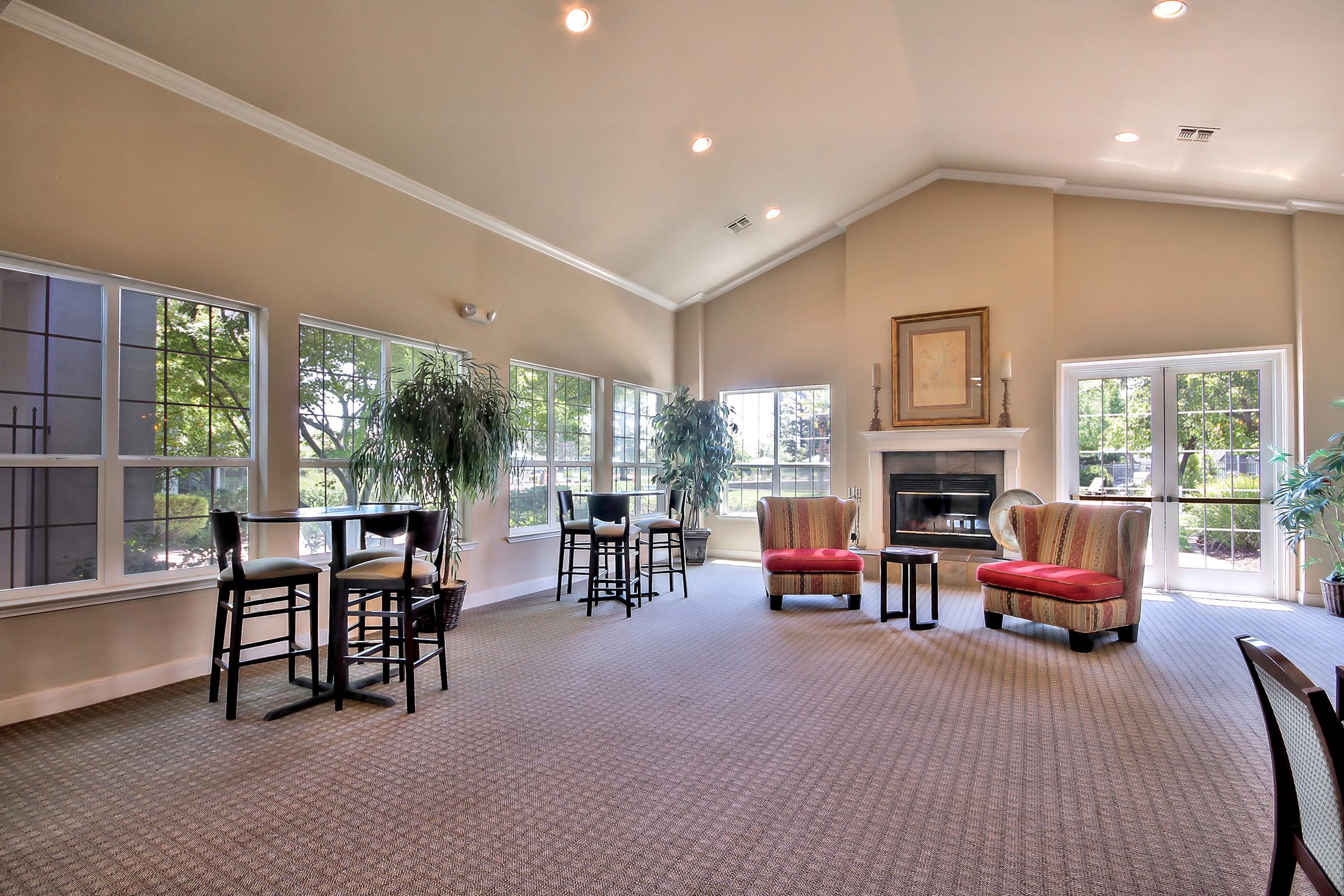Spacious lounge area with large windows providing natural light, featuring several seating options, including high-top tables and armchairs. A cozy fireplace is visible, and decorative plants add a touch of greenery to the environment. The overall atmosphere is inviting and comfortable.