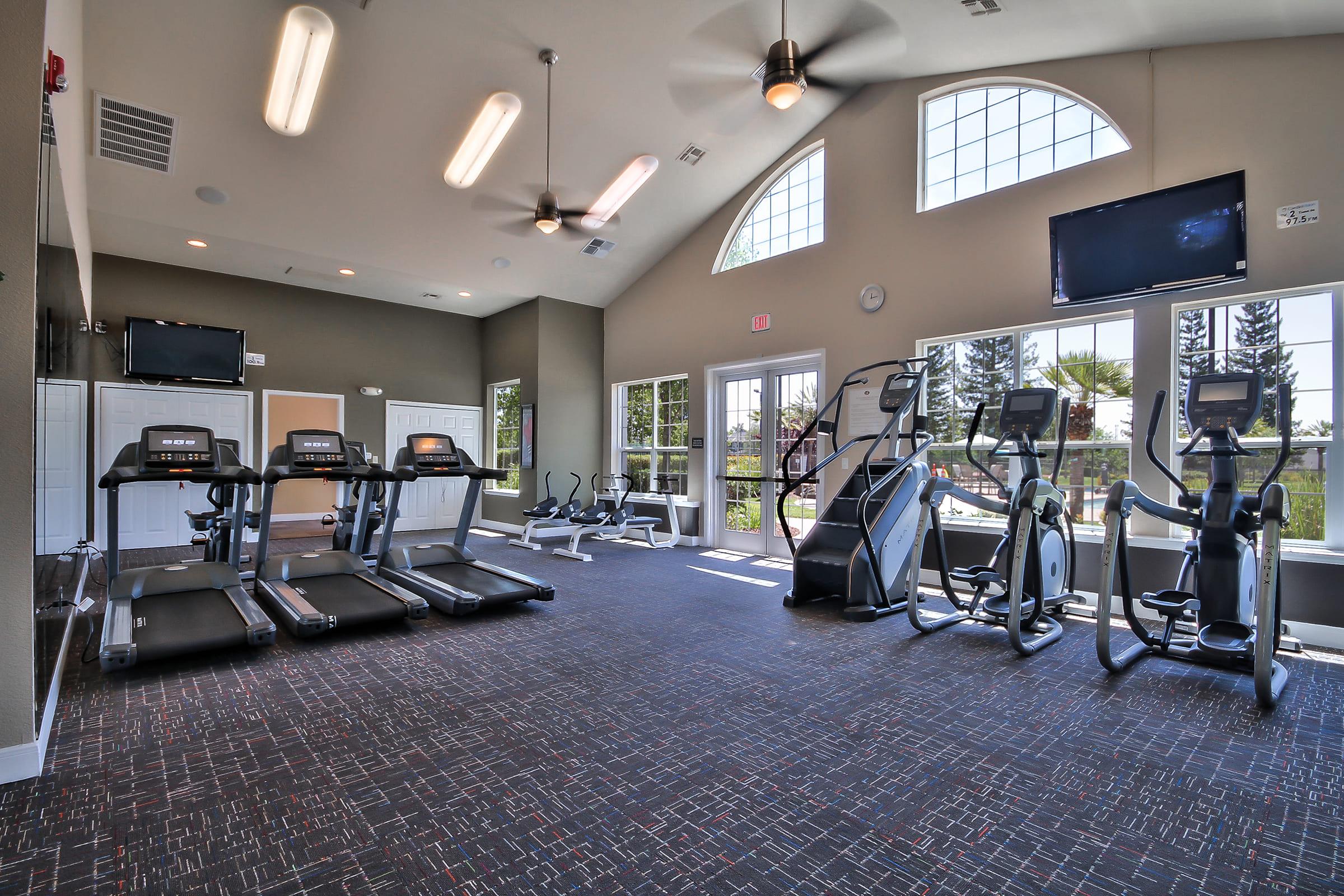 Modern fitness center with treadmills, ellipticals, and a large window allowing natural light. The room features ceiling fans, wall-mounted TVs, and a view of greenery outside. The flooring is a dark carpet with a textured pattern, creating a welcoming and motivating workout environment.