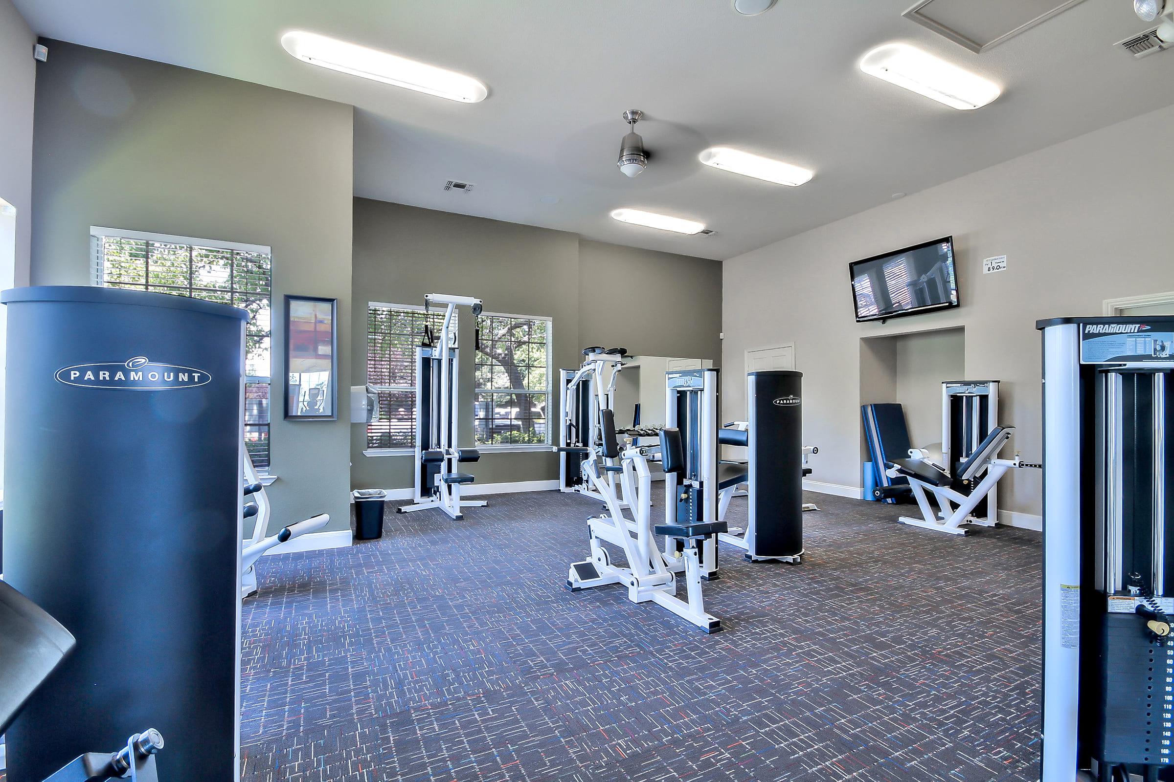 A well-lit gym interior featuring several exercise machines, including strength training equipment. The walls are painted in neutral colors, with large windows allowing natural light. A television is mounted on the wall, and the floor is covered with carpet.