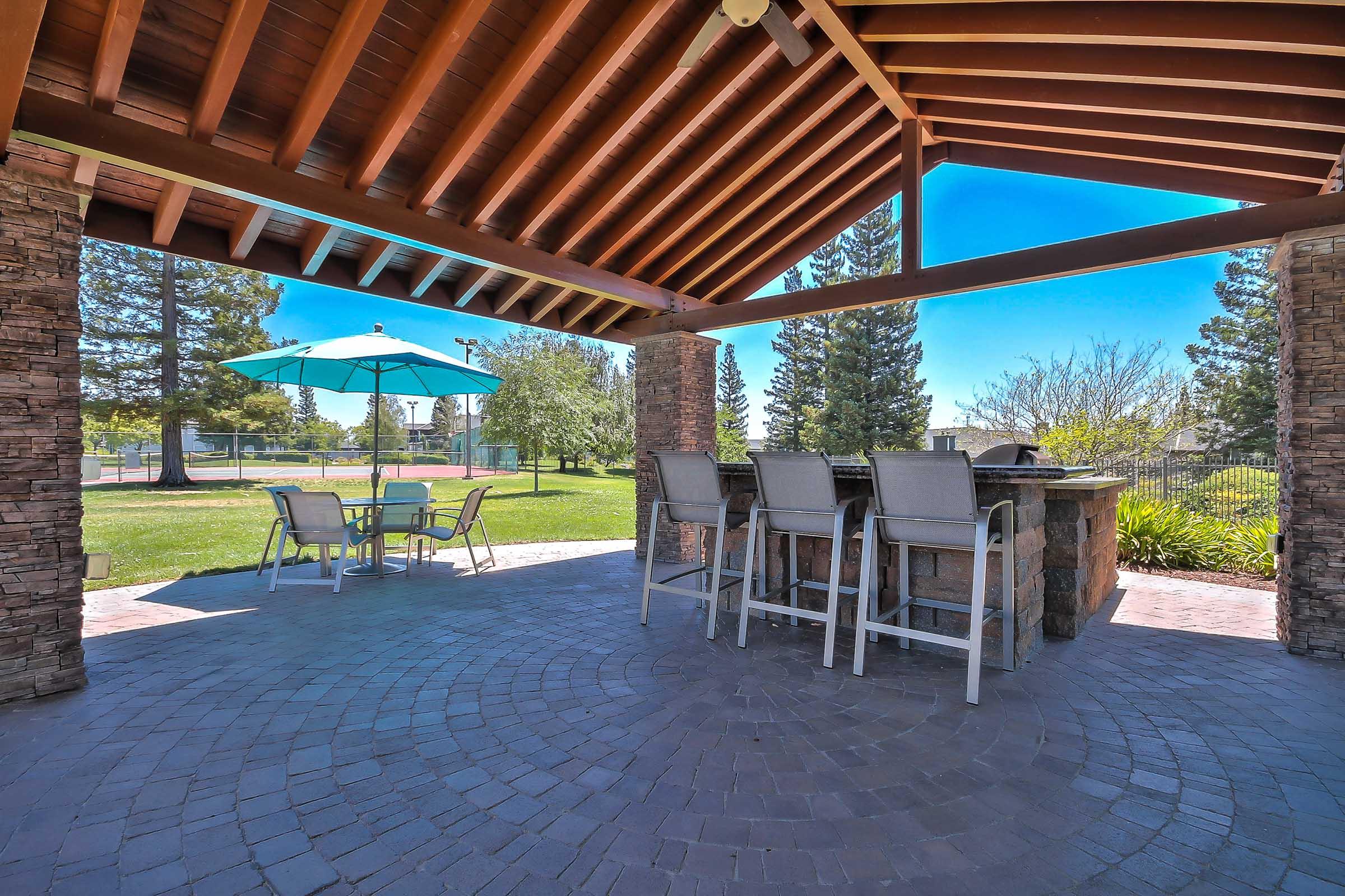 Outdoor patio area featuring a covered seating arrangement with bar stools, a stone bar, and a table with an umbrella. A grassy yard with trees and a sports court can be seen in the background under a clear blue sky.
