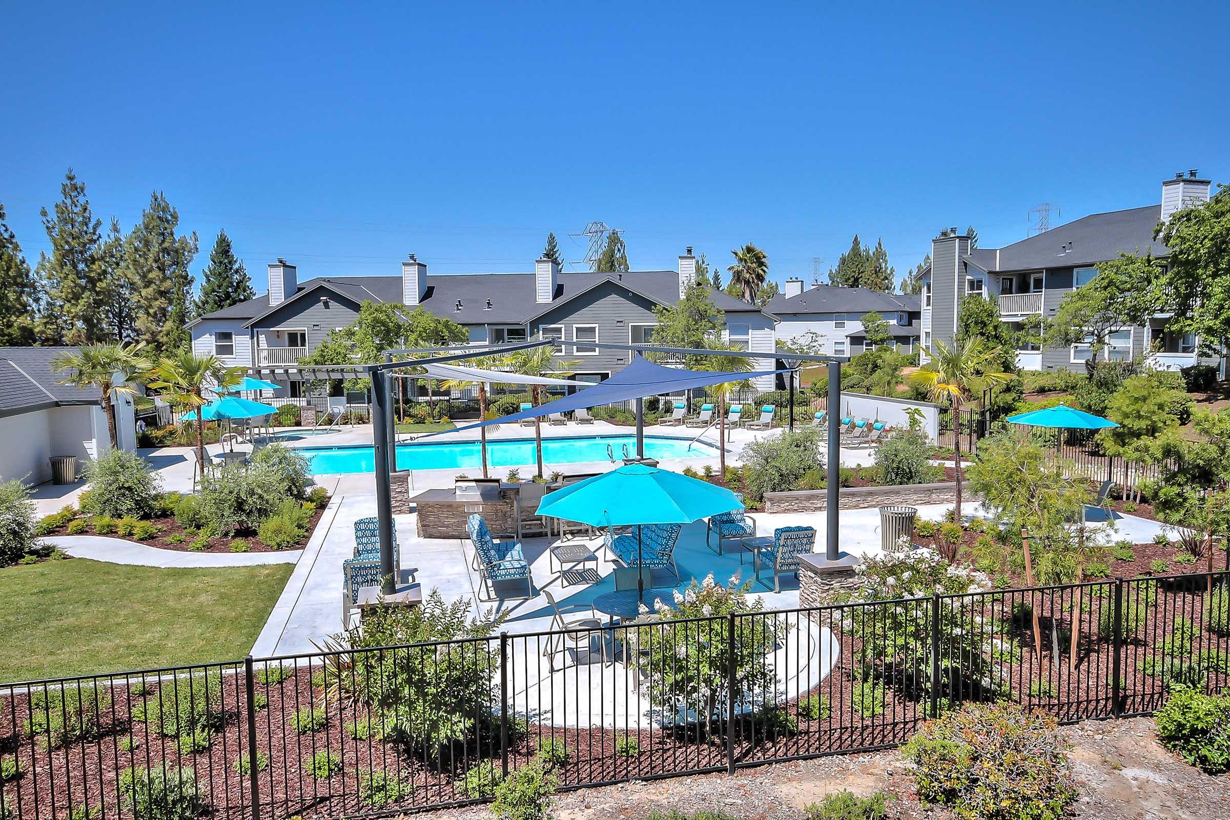 A spacious outdoor area featuring a clear blue swimming pool surrounded by lounge chairs and tables. Bright turquoise umbrellas provide shade, and palm trees and well-maintained greenery enhance the tranquil setting. Modern residential buildings are visible in the background under a clear blue sky.
