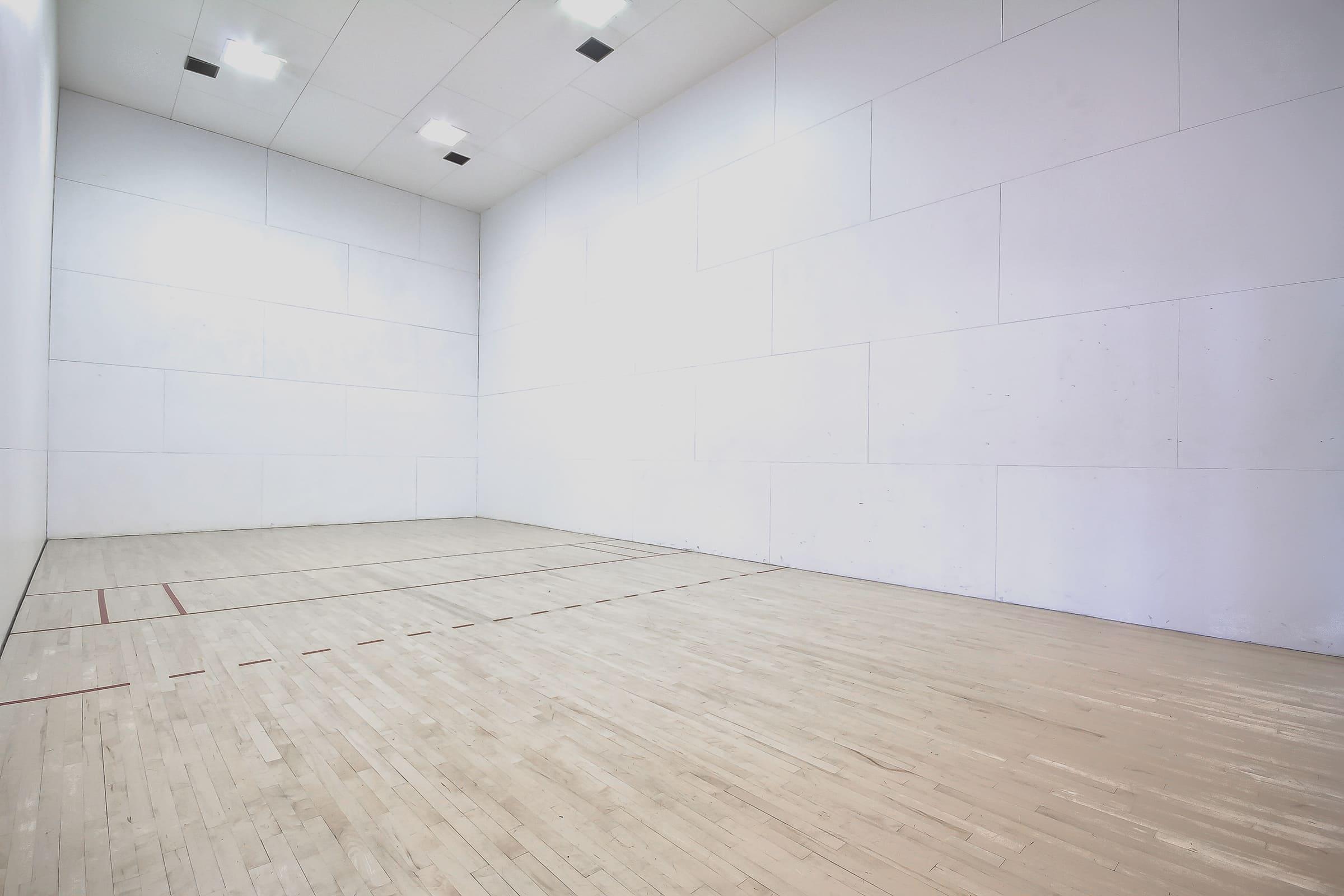 A spacious indoor racquetball court featuring clean white walls and a wooden floor. The court is well-lit, with a high ceiling and red markings on the floor indicating the court boundaries. No furniture or people are present, creating an empty and open atmosphere ideal for playing racquetball.