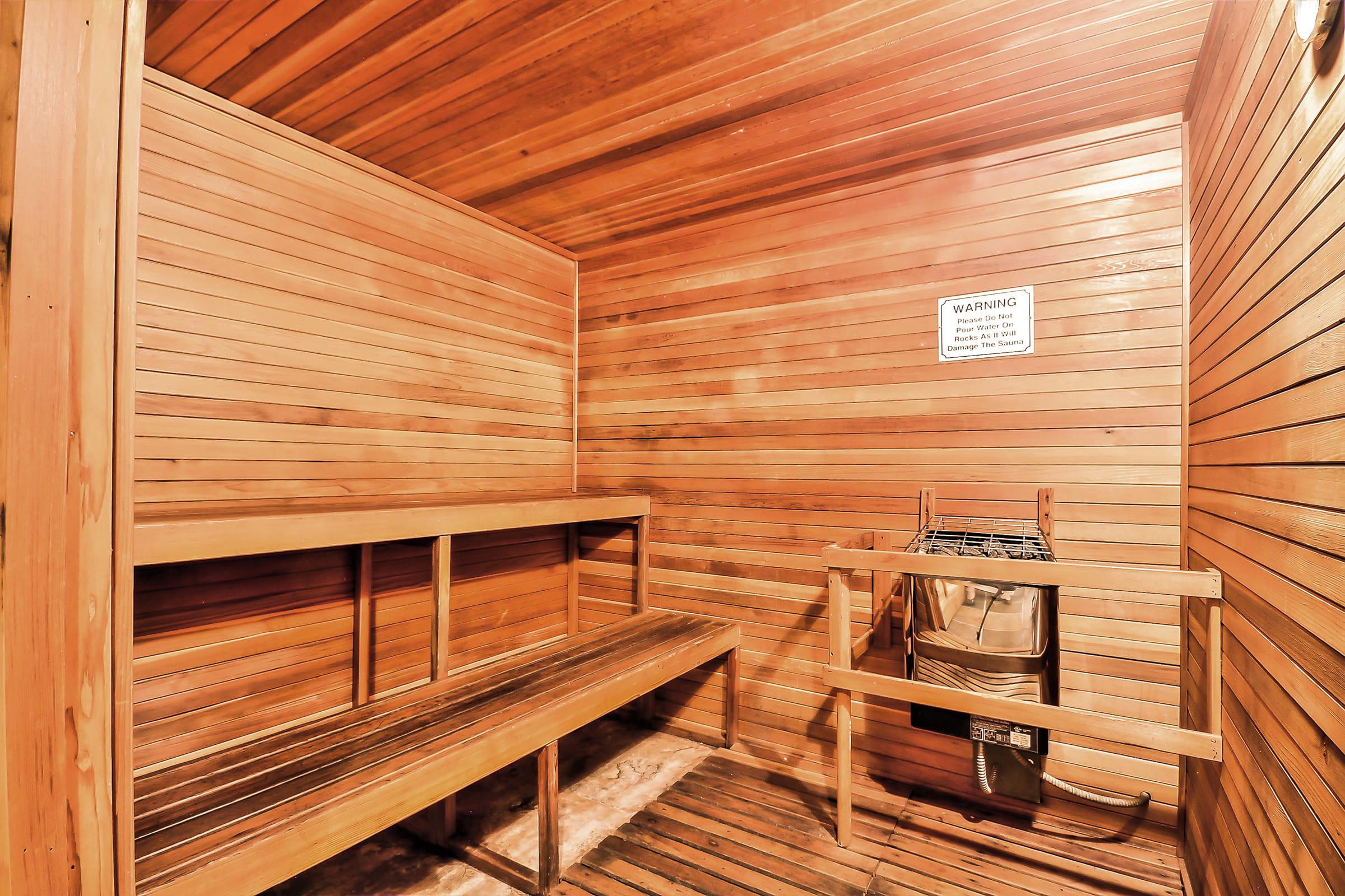 Interior of a wooden sauna featuring two benches, a stove with a water bucket, and a warning sign on the wall. The warm wood paneling creates a cozy atmosphere, inviting relaxation and wellness. The floor is wooden, and the space is well-lit, emphasizing the natural elements of the sauna design.