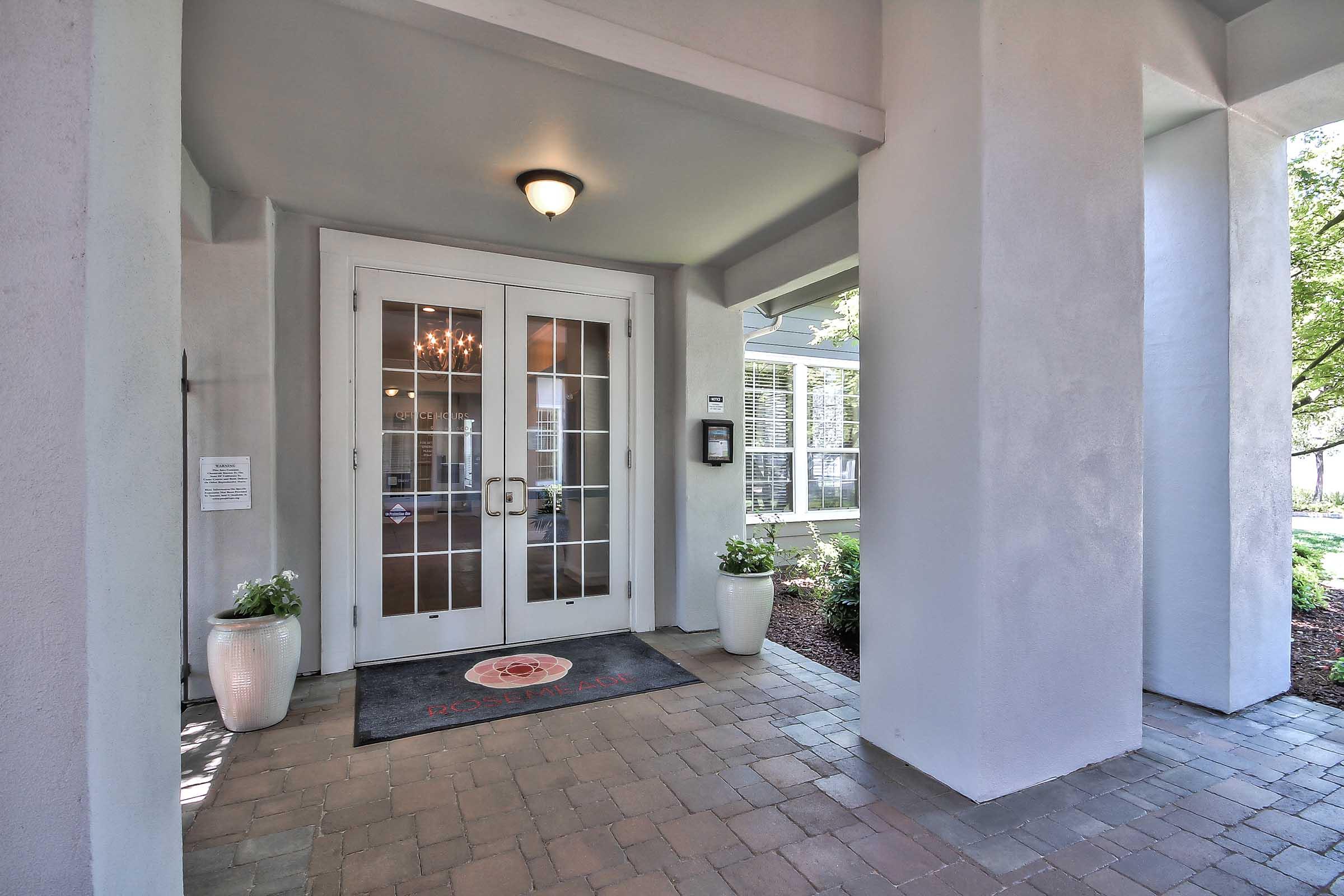 Entrance to a building featuring double glass doors framed by gray columns. Decorative potted plants are placed on either side of the entrance, and a circular logo mat is situated in front. Warm lighting is visible above the doors, creating an inviting atmosphere.