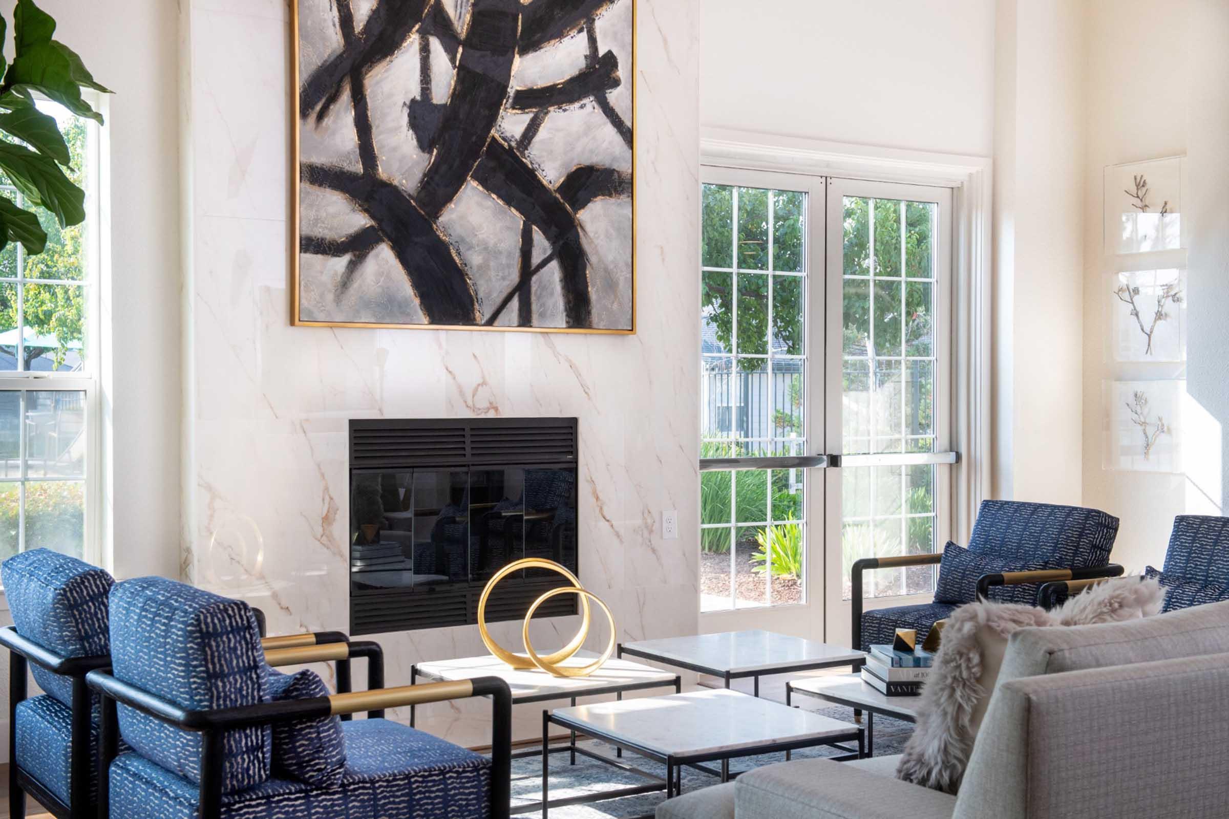 Modern living room featuring a minimalist design with a large abstract painting above a fireplace. Two blue armchairs with wooden frames and a plush beige sofa are arranged around a coffee table. Natural light streams in through large windows, with greenery visible outside.