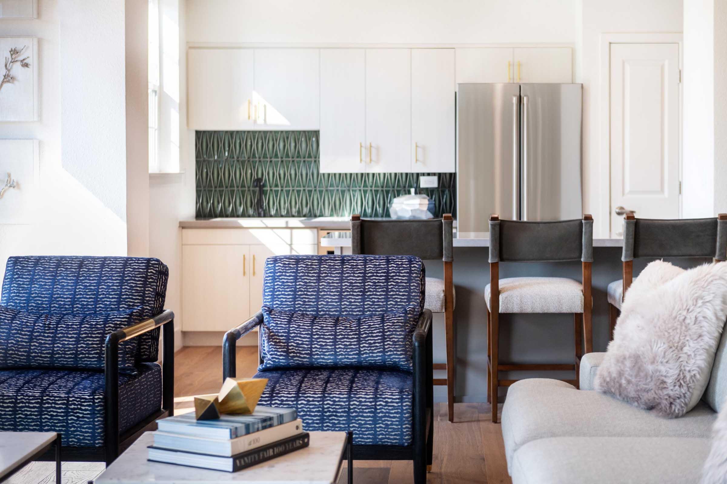 A modern and stylish kitchen and living area featuring two patterned blue chairs and a light gray sofa. The kitchen has white cabinets, a green patterned backsplash, and stainless steel appliances. A small table with books sits in front of the chairs, enhancing the cozy atmosphere.