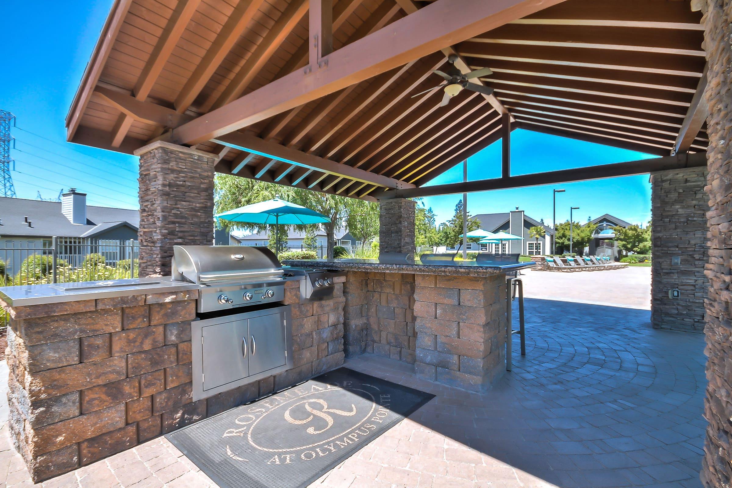 An outdoor BBQ area featuring a stone grill station under a wooden gazebo, surrounded by lush landscaping and a pool area in the background. The setting is bright and inviting, ideal for outdoor cooking and gatherings.