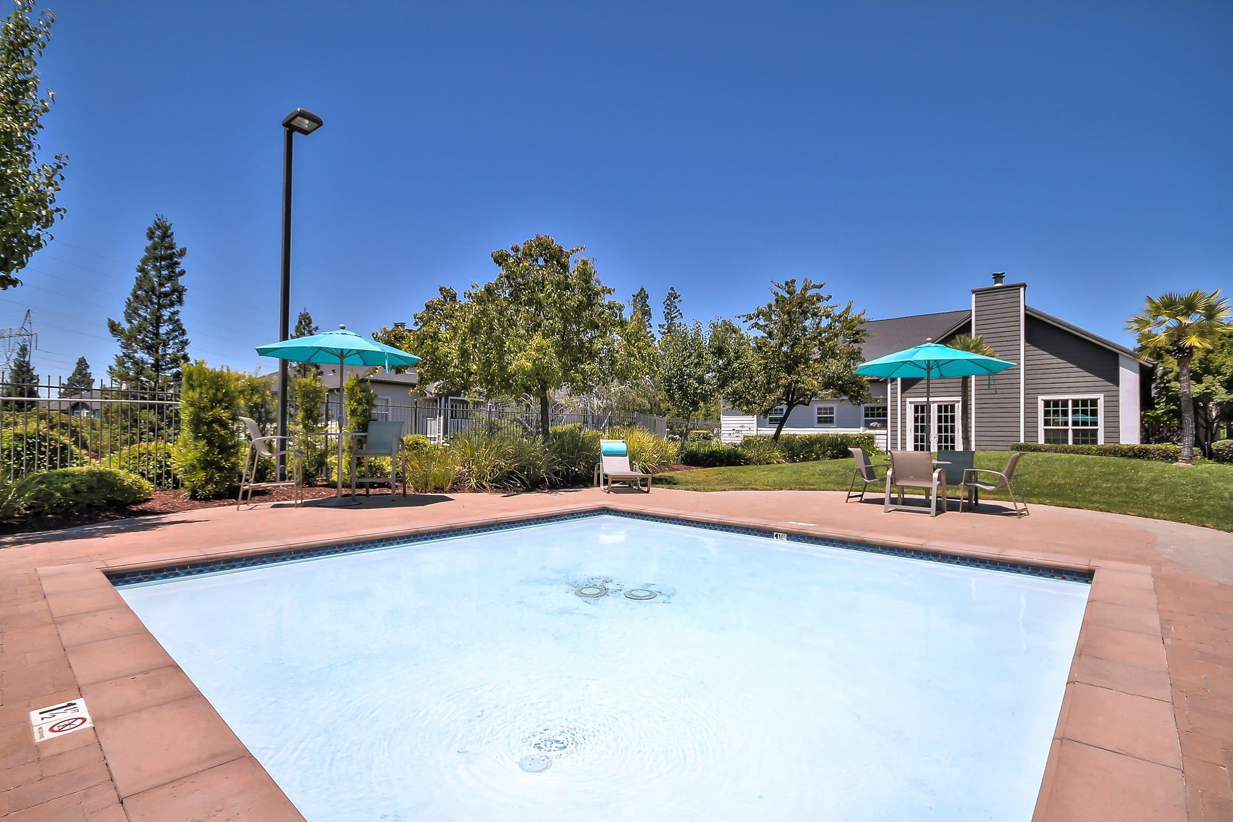 A clear swimming pool with a light blue surface, surrounded by green grass and trees. There are two umbrellas providing shade over lounge chairs nearby. In the background, a modern building is partially visible under a bright blue sky.