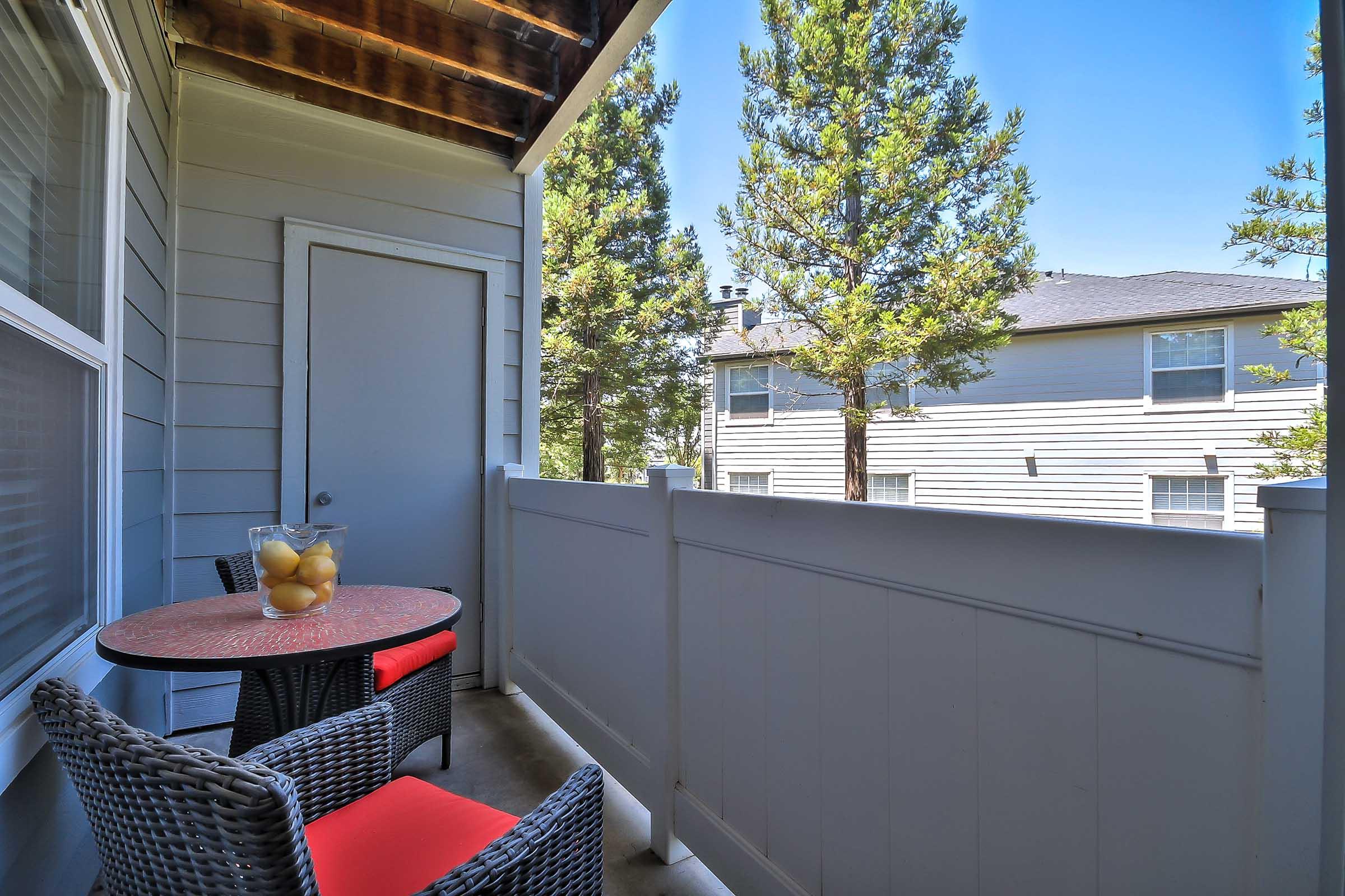 A cozy balcony with a small round table and two chairs, featuring a bowl of fruit on the table. The area is surrounded by a white fence and shows trees and neighboring buildings in the background under a clear blue sky.