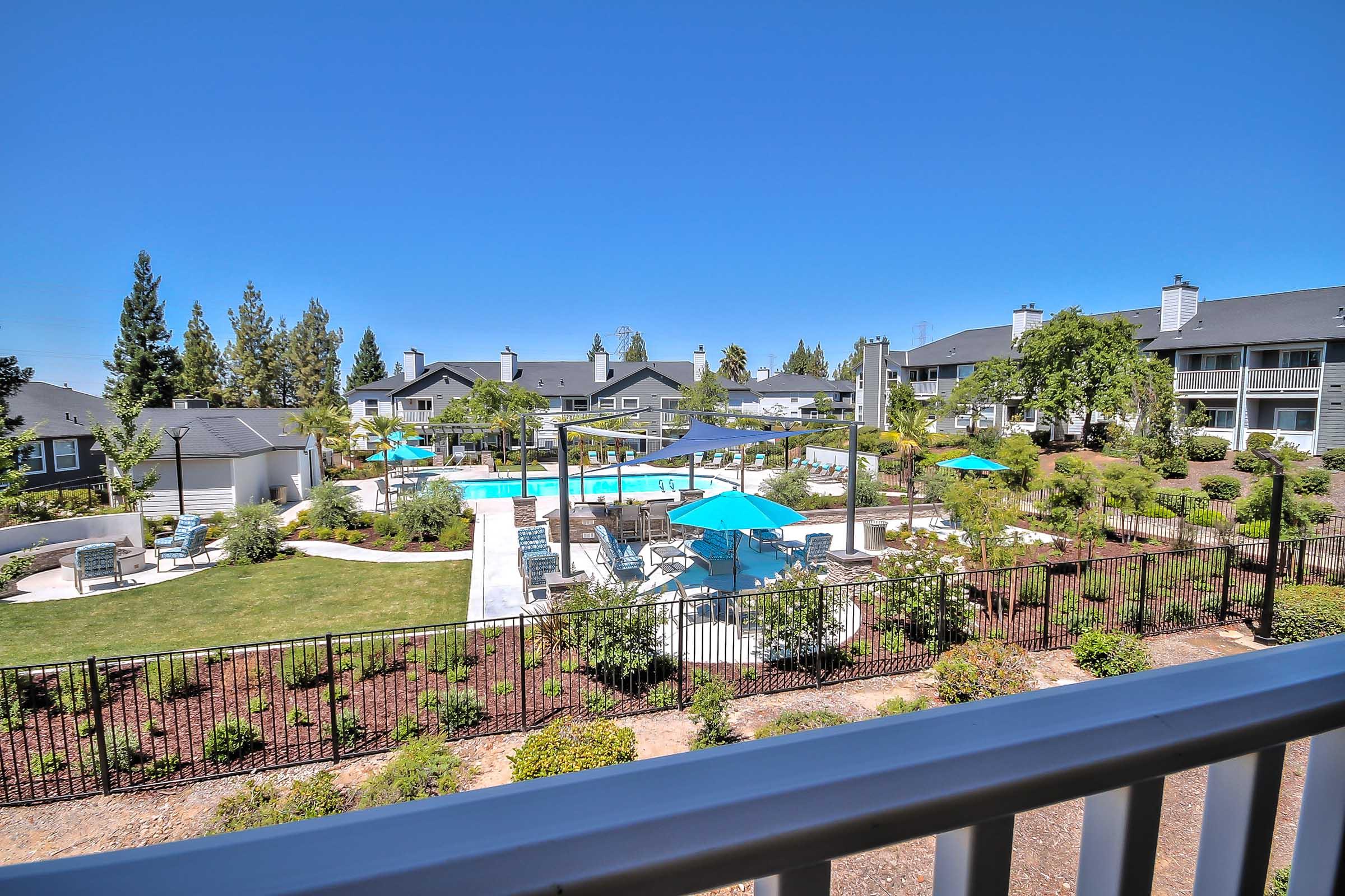 A scenic view of a residential pool area featuring a large swimming pool surrounded by lounge chairs and umbrellas. The landscape includes well-maintained greenery and trees, with several buildings in the background, showcasing a bright blue sky.