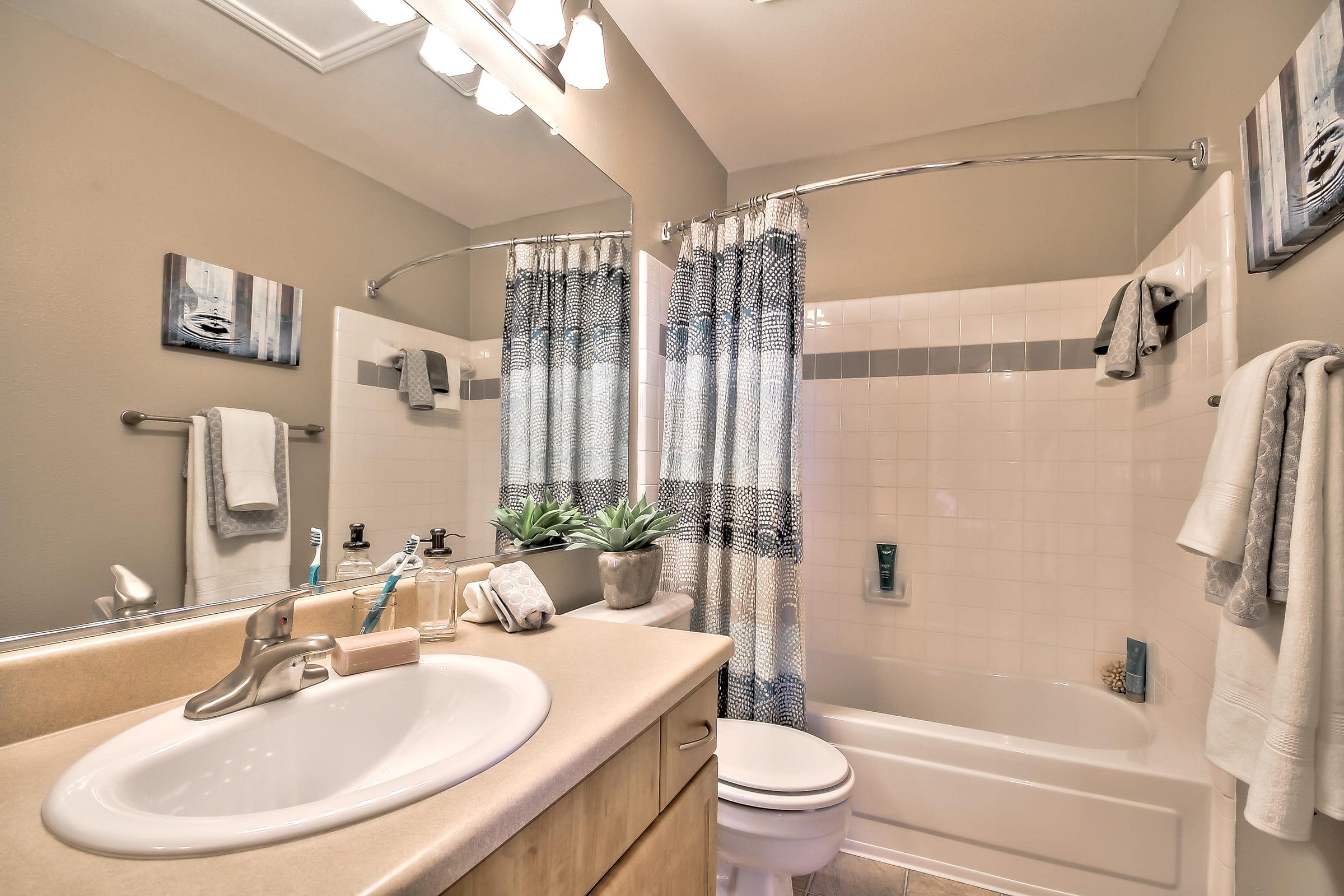 A modern bathroom featuring a bathtub with a curved shower rod, light-colored walls, and a beige countertop. The space has decorative elements like a plant, neatly arranged towels, and framed artwork on the walls, contributing to a clean and organized look.