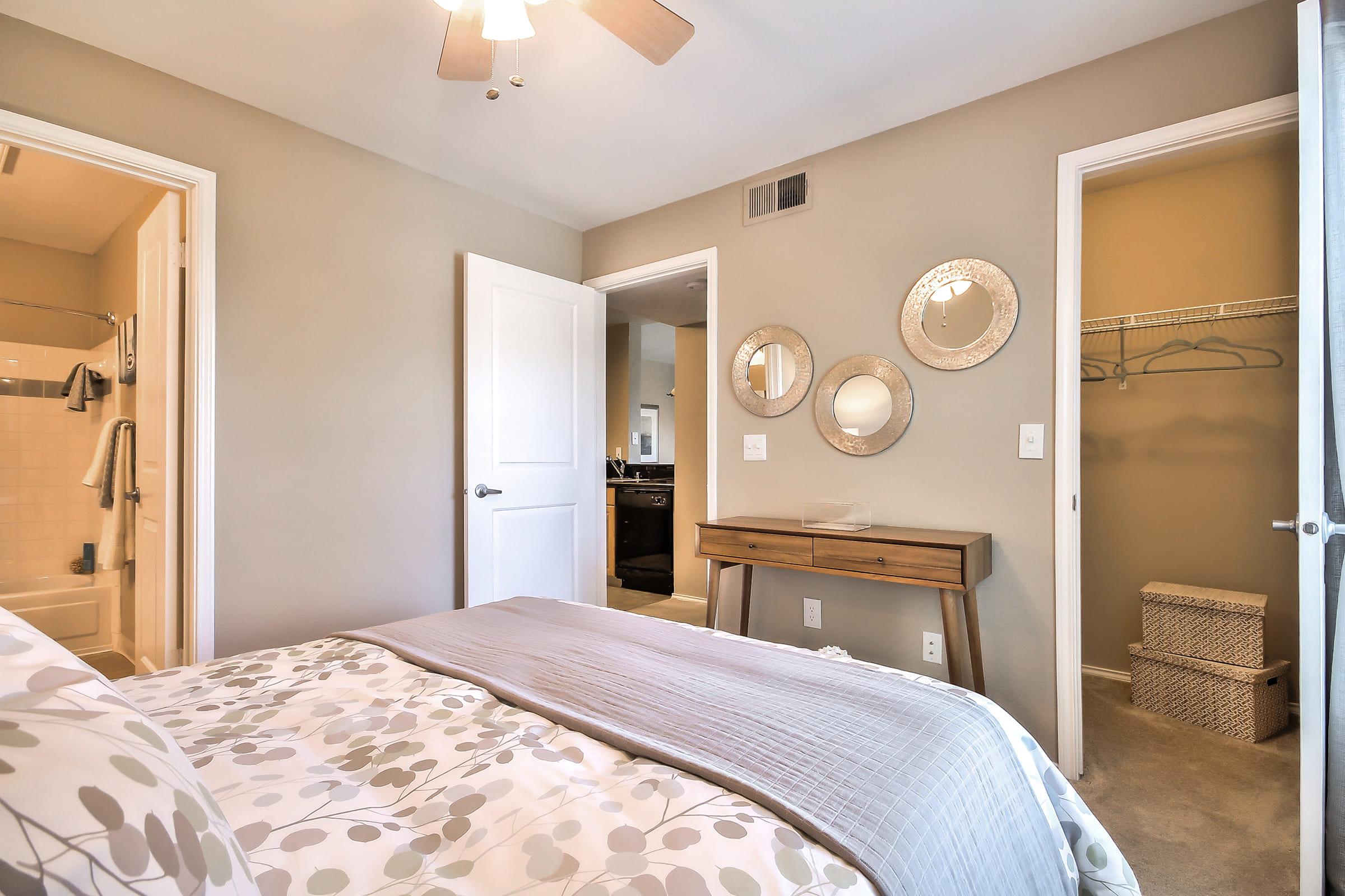 A well-lit bedroom featuring a bed with a light-colored duvet. There's a minimalist wooden side table and mirrors on the wall. Two open doors lead to a bathroom and a closet. The walls are painted in a soft beige, adding warmth to the space