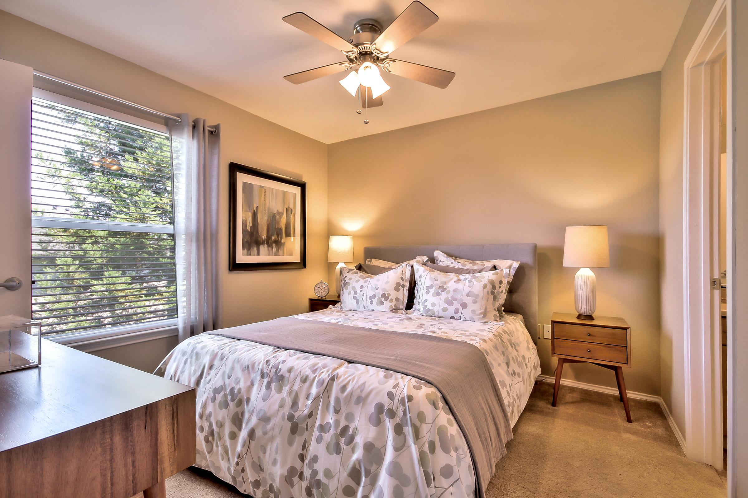 A cozy bedroom with a queen-sized bed featuring a gray and white patterned duvet. There are two decorative pillows on the bed. A wooden nightstand holds a lamp, and a large framed artwork hangs on the wall. Natural light enters through a window with blinds, and a ceiling fan is mounted above.