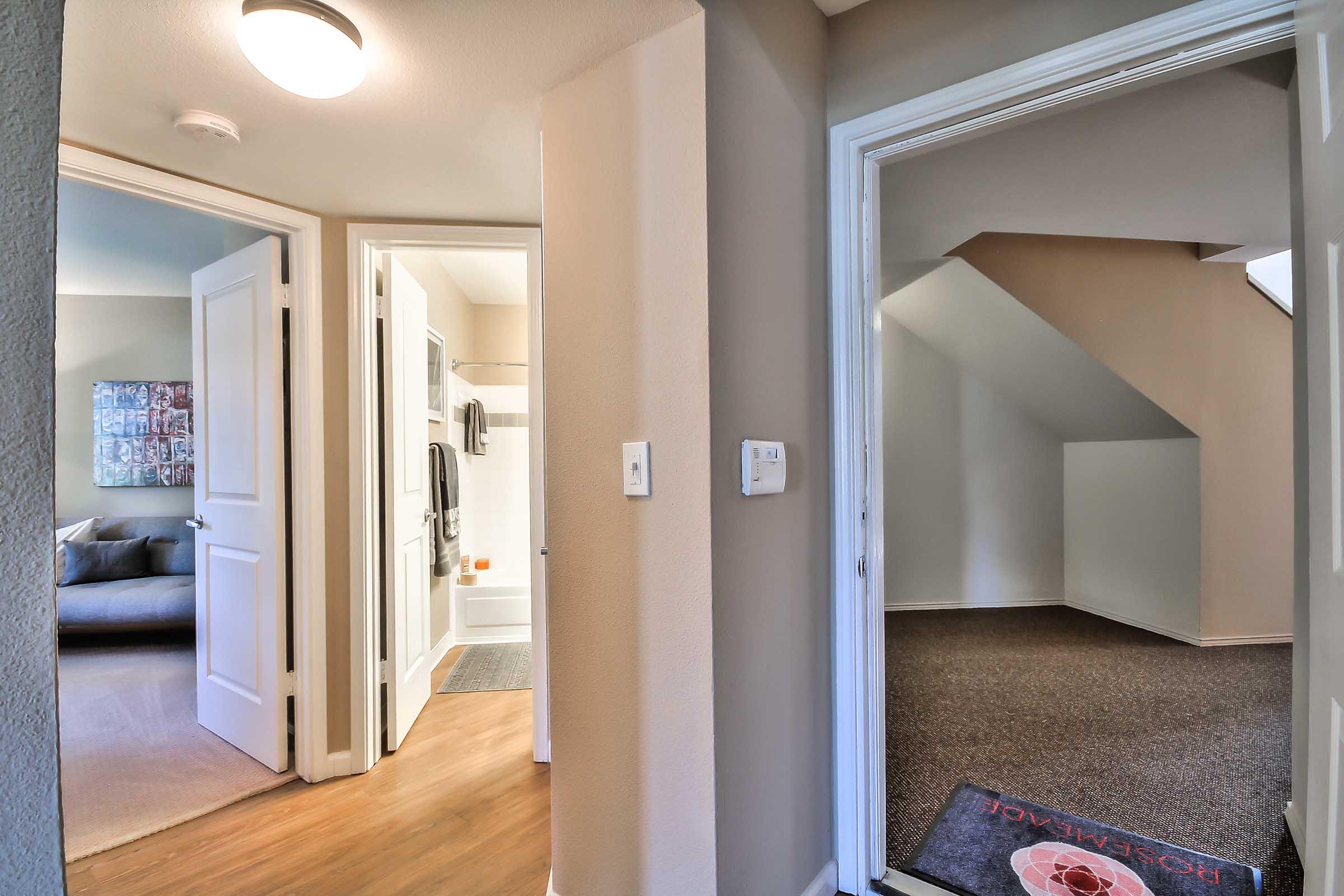 View of an interior hallway with two open doors. The left door leads to a bathroom with white walls and fixtures, while the right door opens to an empty room with carpeted flooring. A modular sofa can be seen in the distance through the left doorway.