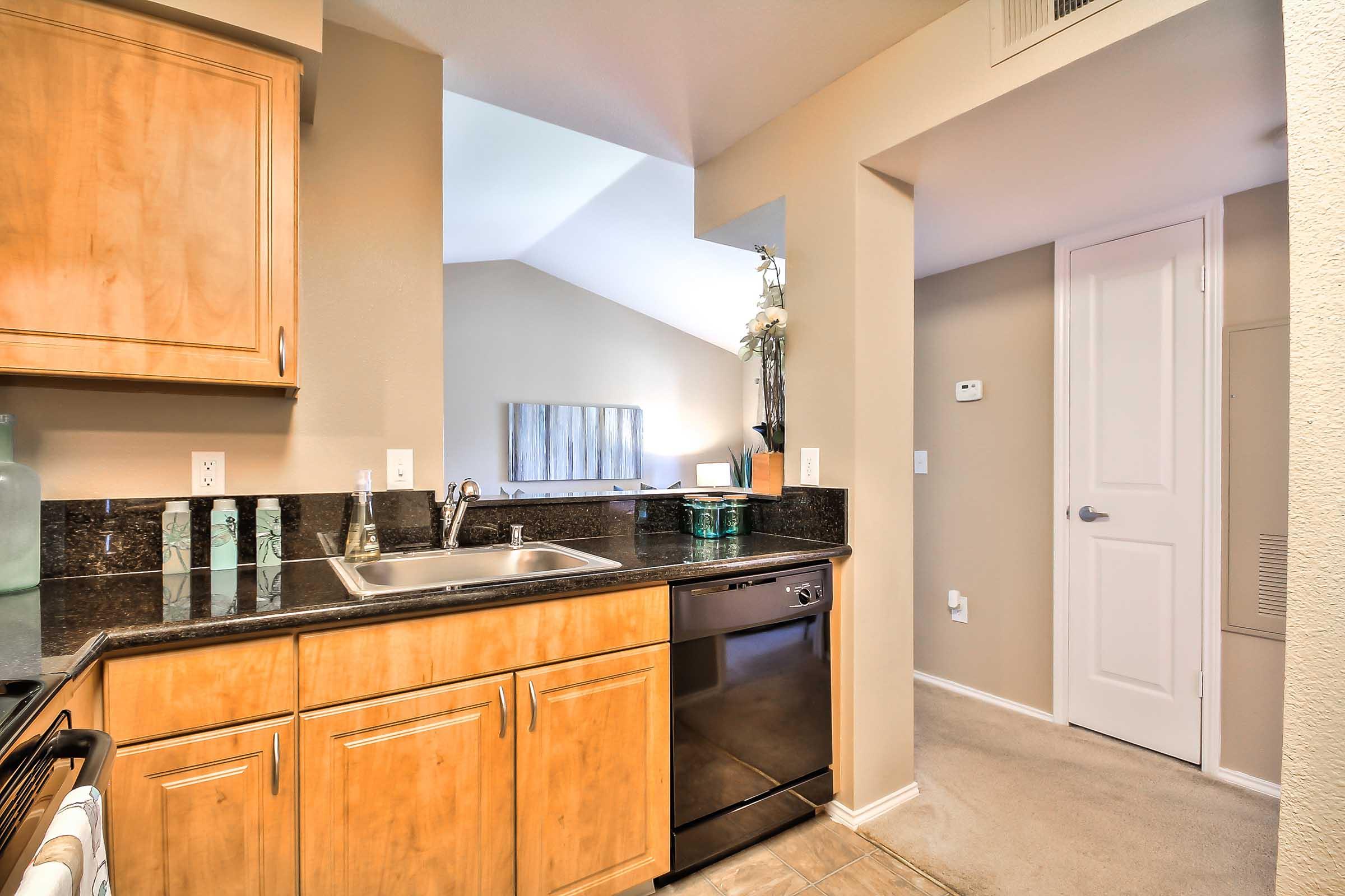 : A modern kitchen featuring wooden cabinetry, a stainless steel sink, and a dishwasher. The countertop is dark granite, and the space has beige walls. An adjacent area shows a light-filled room with a vaulted ceiling and a door leading to another room.
