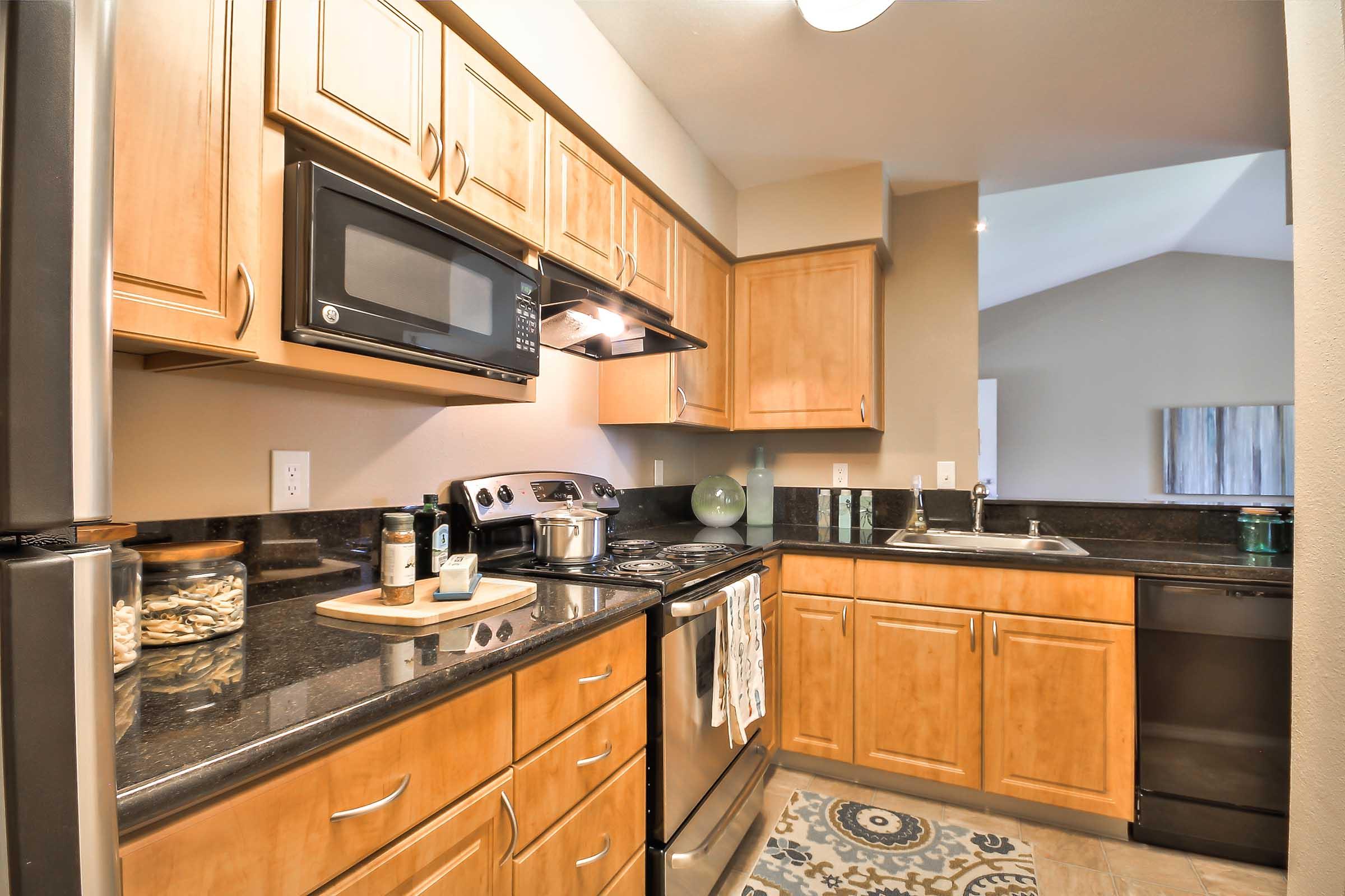 Modern kitchen featuring wooden cabinetry, stainless steel appliances, and black granite countertops. Includes a microwave, stove, and sink with a decorative rug on the floor. Small decorative items and a bowl of snacks are visible on the countertop. The space is well-lit, showcasing an open layout.