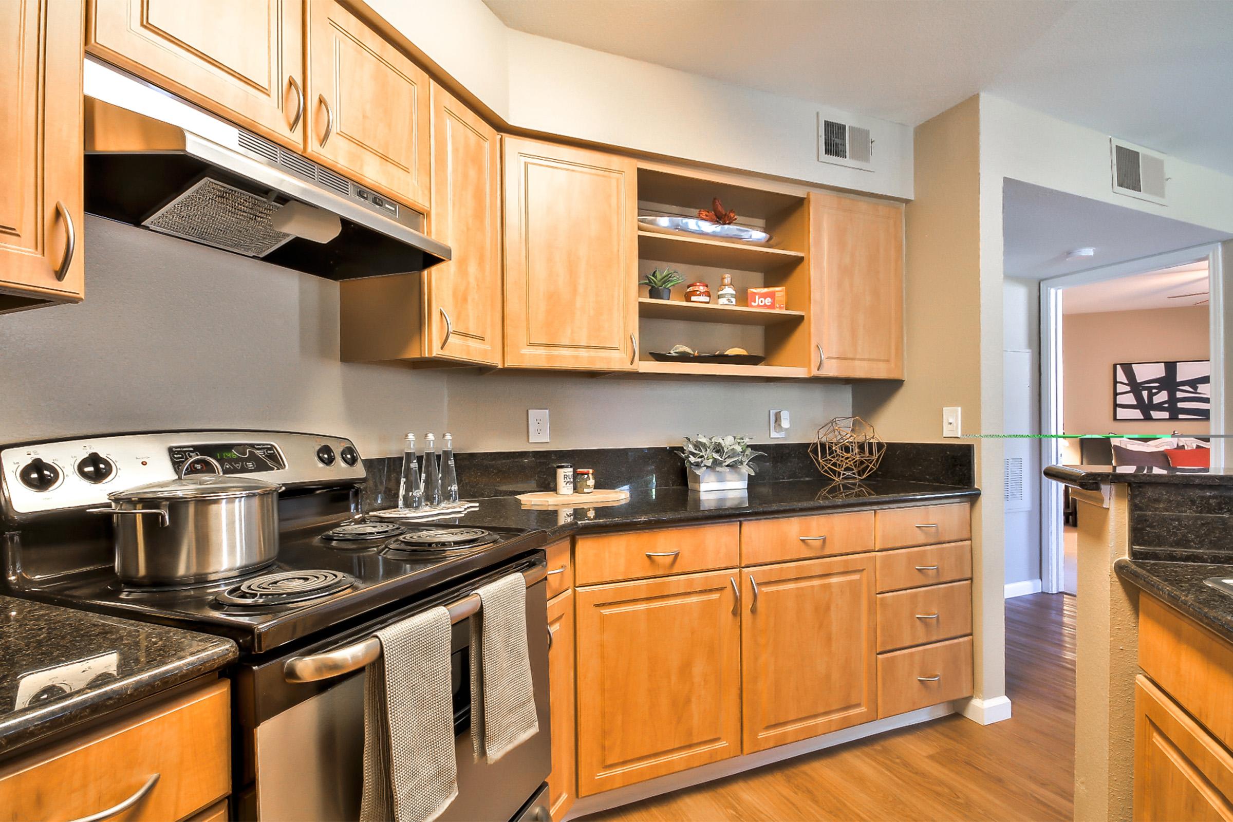 A modern kitchen features wooden cabinets, a black granite countertop, and stainless steel appliances. There is a stove, a pot on the burner, and decorative items on an open shelf. The space is well-lit and organized, showcasing a clean and inviting cooking environment.