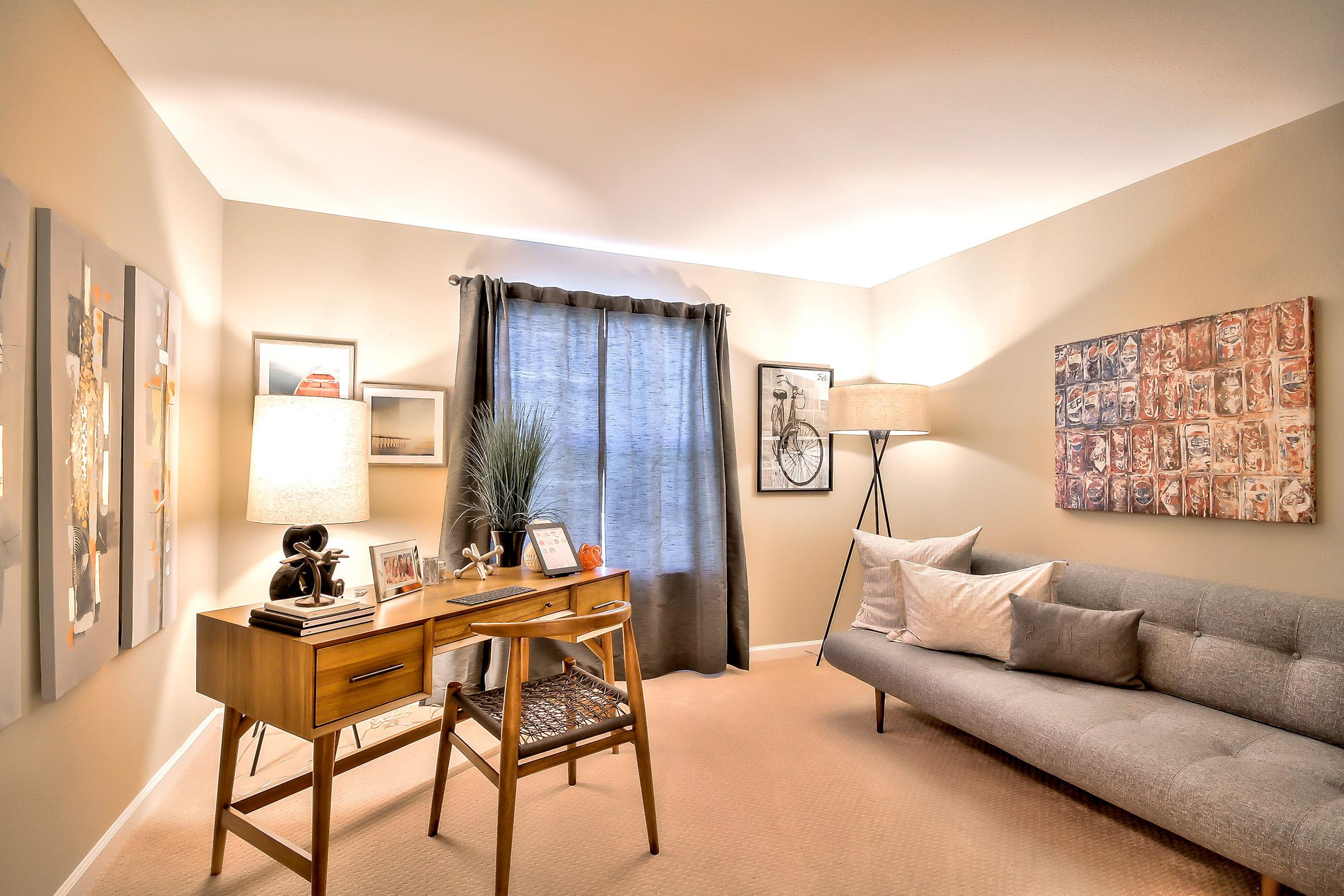 A cozy modern office space featuring a sleek gray sofa, a wooden desk with decorative items, and art on the walls. A lamp illuminates the room, which has neutral-colored walls and a window with dark curtains. The overall atmosphere is inviting and stylish.