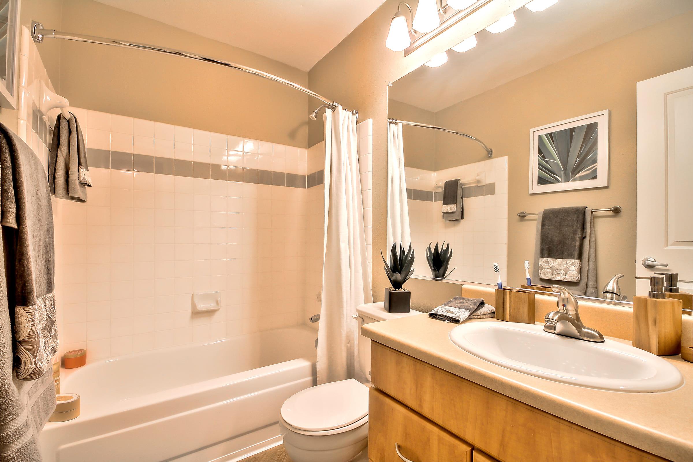 A well-lit bathroom featuring a white bathtub with a shower curtain, a toilet, and a sink with wooden cabinetry. Decorative elements include potted plants and neatly arranged towels. The walls are painted in a neutral color, creating a clean and inviting atmosphere.