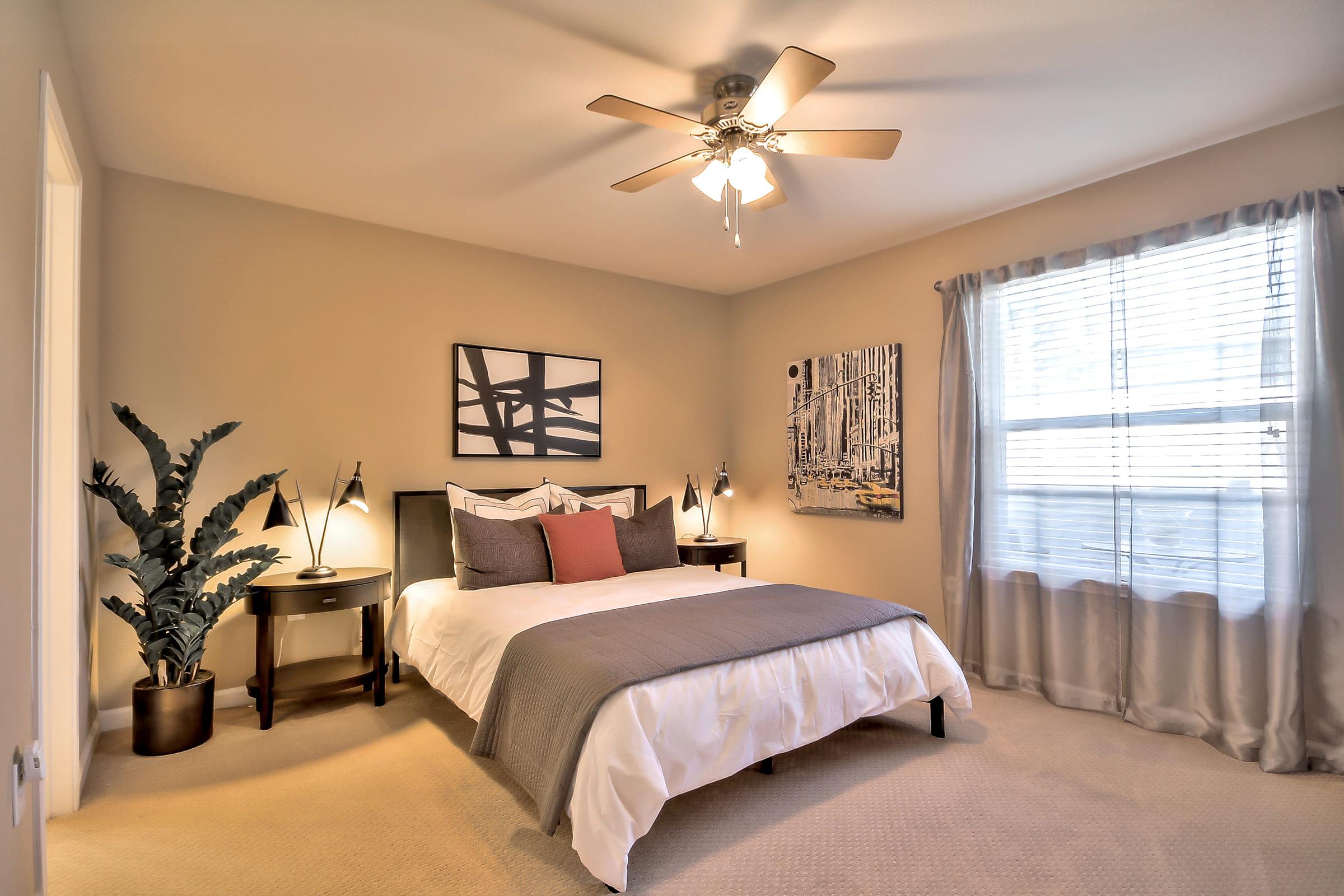 A cozy bedroom featuring a bed with gray and white bedding, two bedside tables with lamps, a large window with sheer curtains, and a decorative plant. The walls are painted in a neutral tone, and artwork is hung above the bed, adding a modern touch to the space. A ceiling fan is also visible.