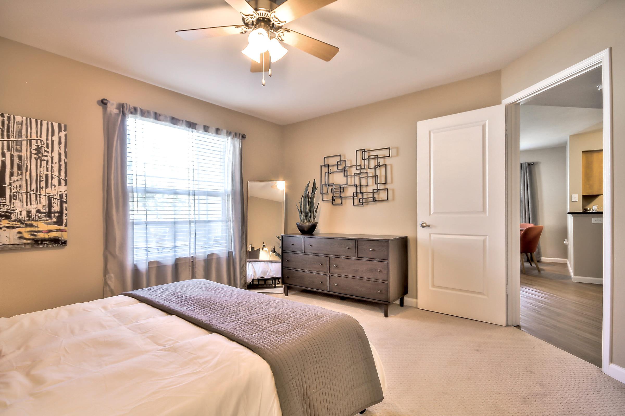 A cozy bedroom featuring a neatly made bed with a gray blanket, a ceiling fan, and large windows with sheer curtains letting in natural light. There's a dark wooden dresser and a wall-mounted decorative art piece. A mirror and indoor plant add a touch of elegance, complementing the neutral colored walls.