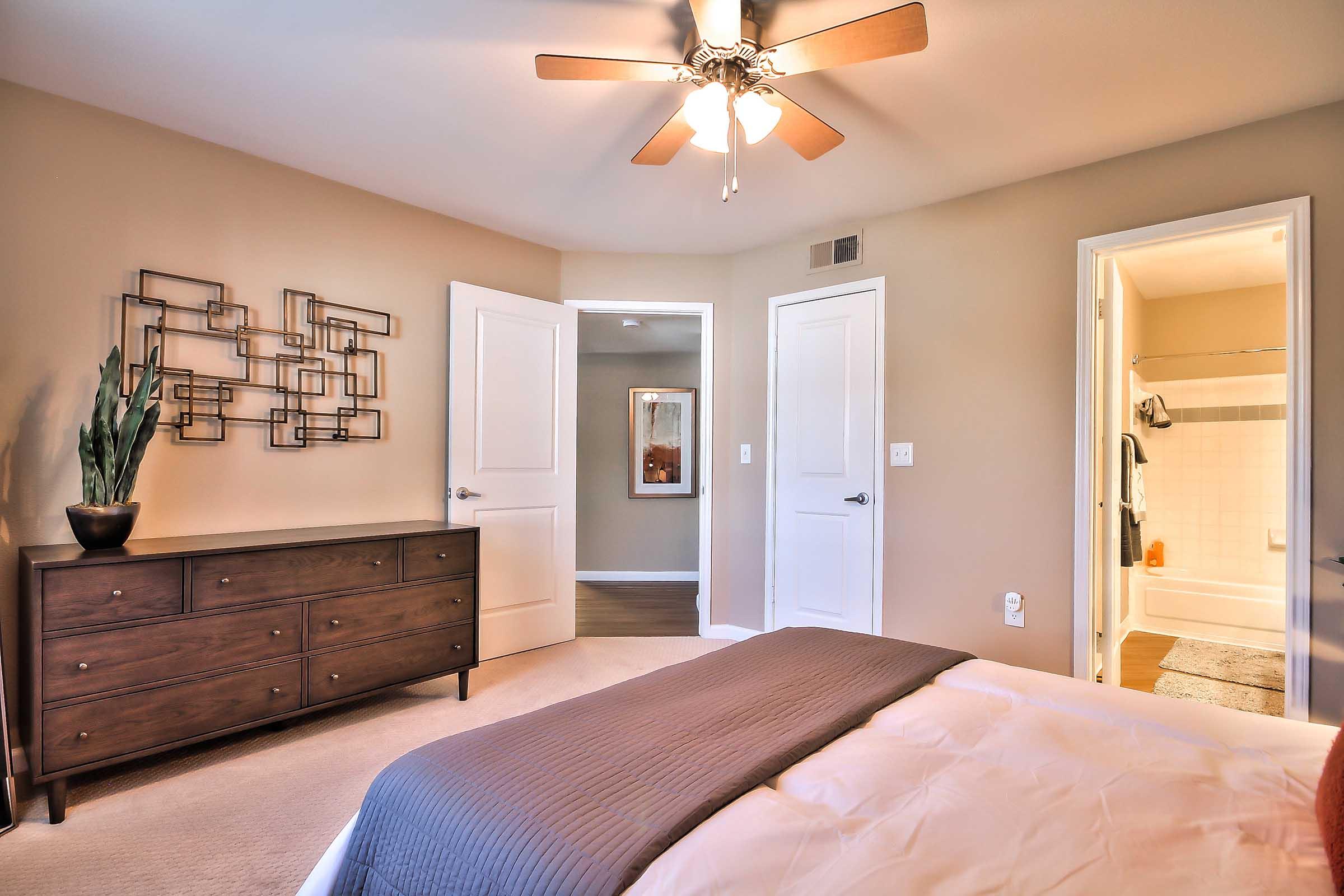 A cozy bedroom featuring a ceiling fan, a large window, and a wooden dresser. The walls are painted in a warm beige tone, and there are two doors leading to other areas of the home. A potted plant sits on the dresser, and a bathroom is visible through one of the doors. The bedding is neatly arranged on the bed.