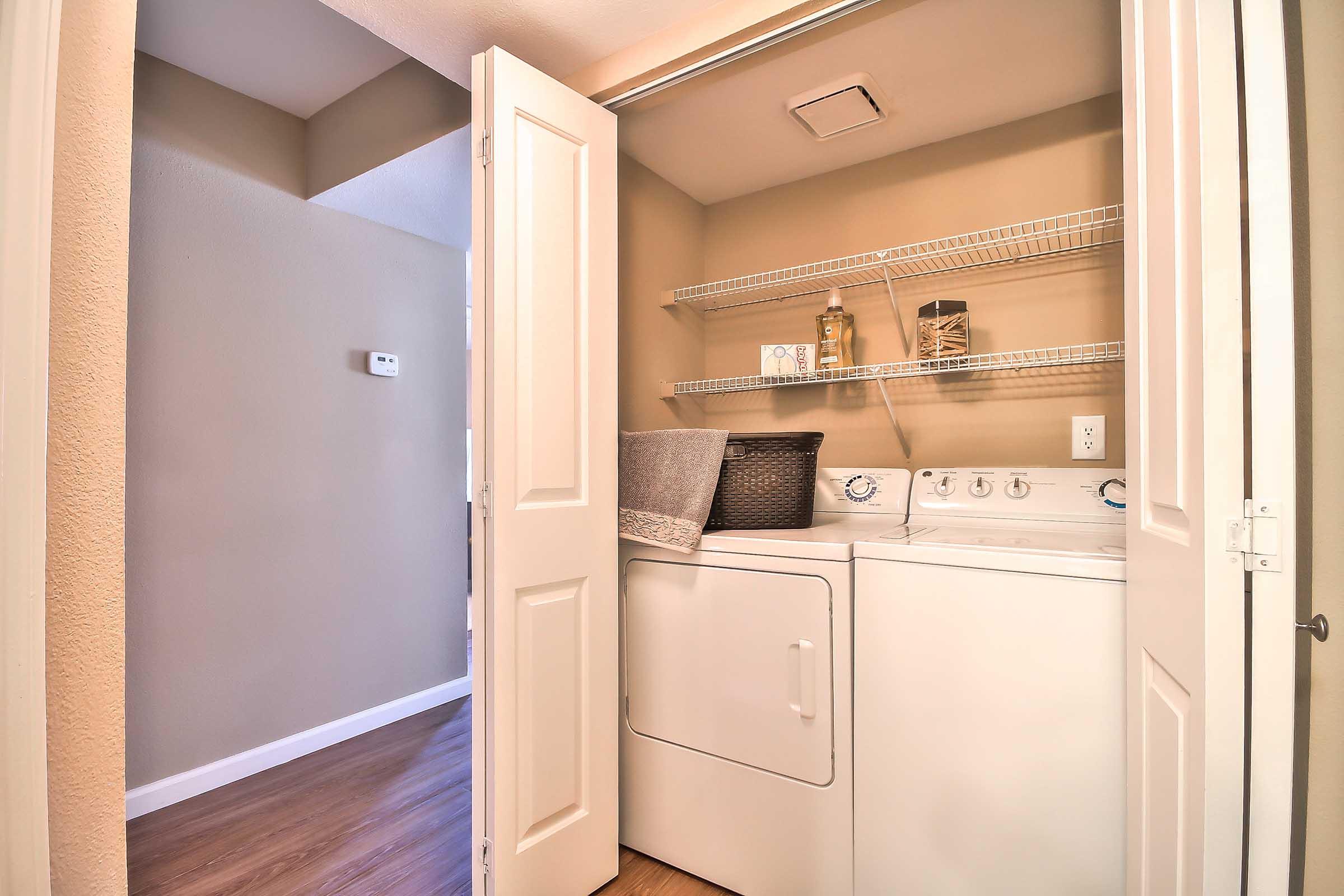 A laundry area featuring a stacked washer and dryer, situated in a small room. Shelving above holds various items, and a dark basket sits on the floor next to a folded grey towel. The walls are painted a light color, and a doorway leads to another room.