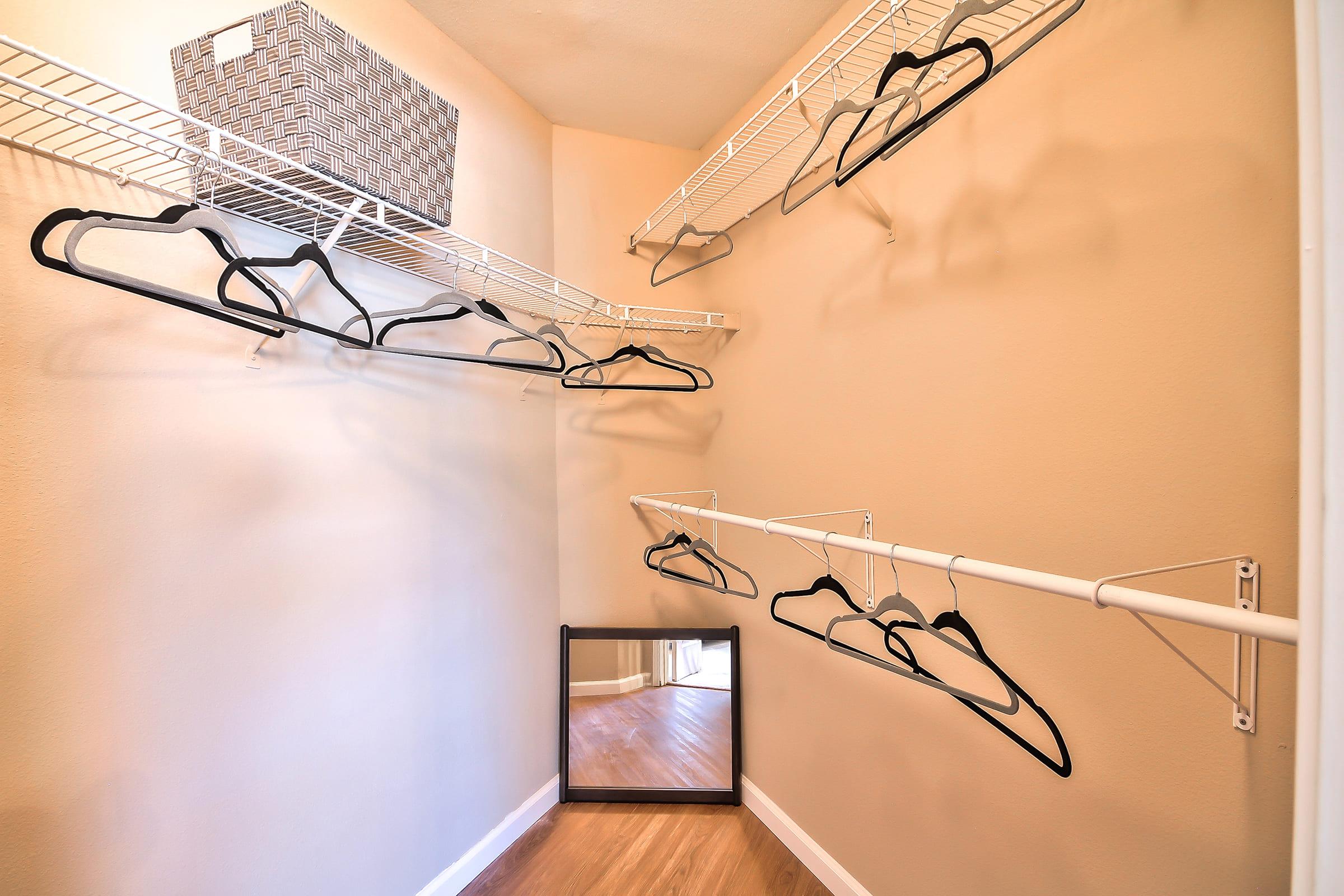 A neatly organized closet with wire shelving, multiple black hangers hanging from the shelves, and a small mirror leaning against the wall. A wicker basket is also on the top shelf, and the walls are painted a soft beige, complementing the wooden floor.