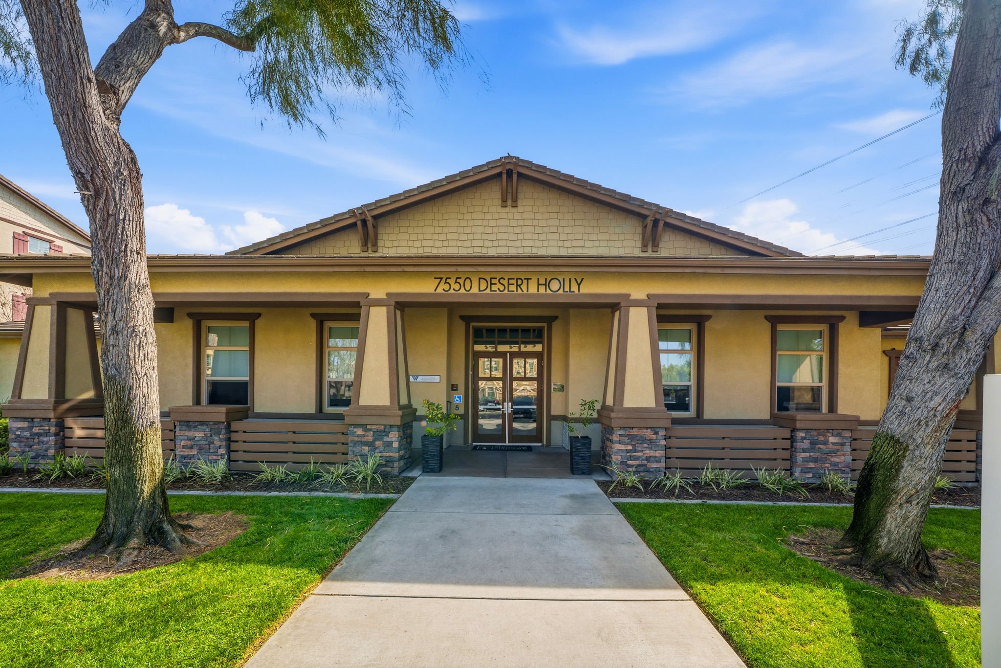 A single-story building with a welcoming facade, featuring a light brown exterior, stone accents, and a pathway leading to a central entrance. The address "7550 DESERT HOLLY" is displayed above the door. Flanked by two trees, the landscaped area includes green grass and shrubs, under a clear blue sky.