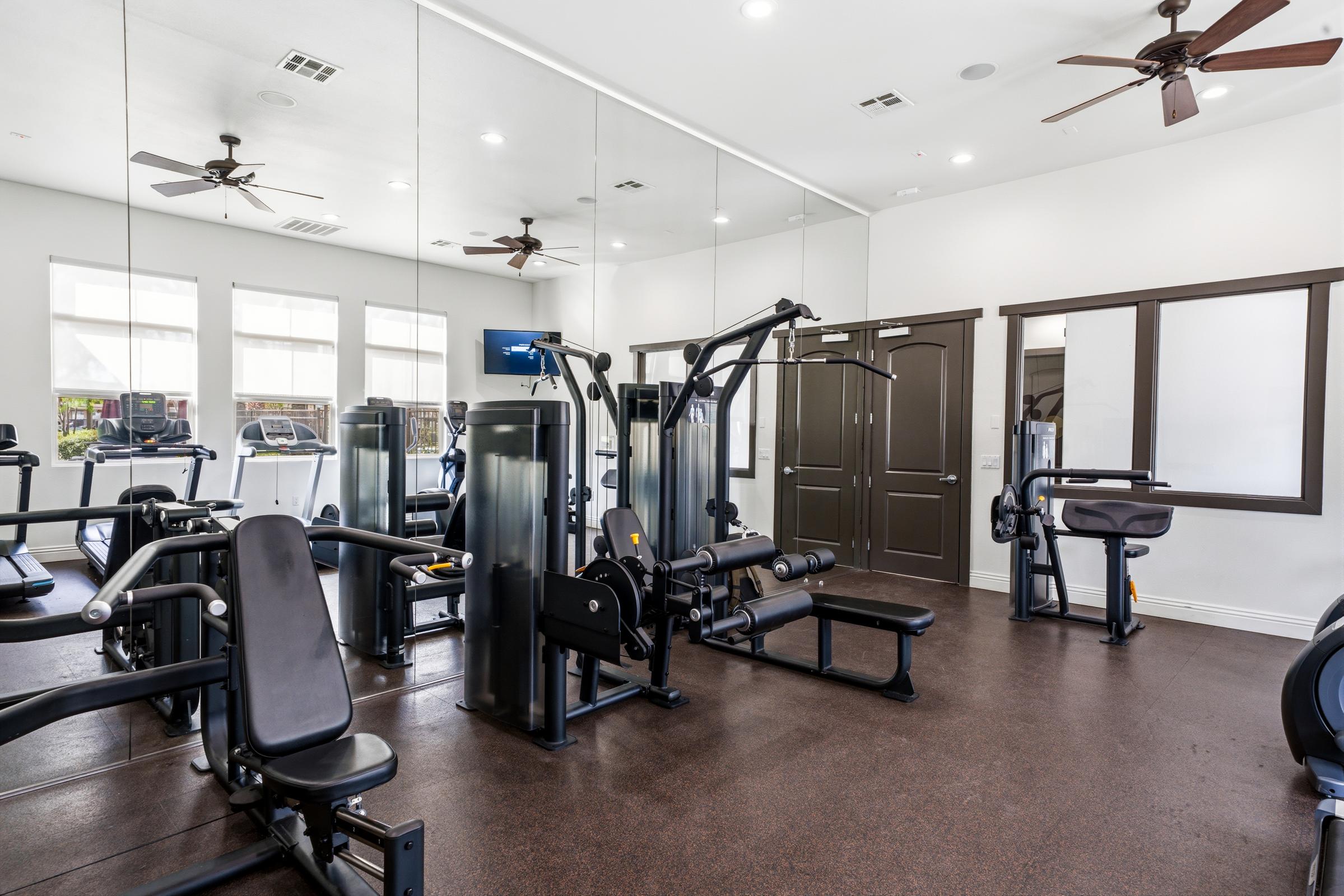 A well-equipped gym interior featuring various exercise machines, including weightlifting and cardio equipment. The space has large mirrors on the walls, ceiling fans for ventilation, and natural light coming from windows. It appears clean and organized, suitable for workouts.