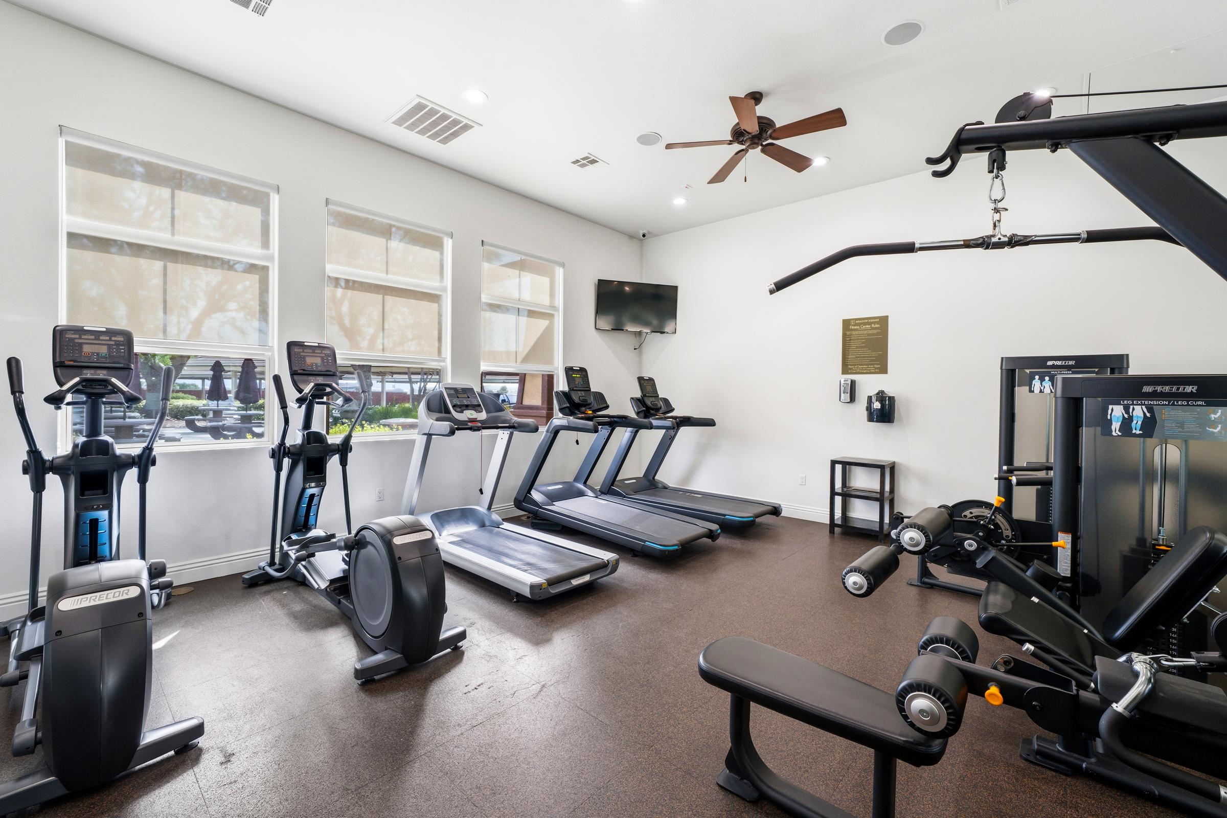 A modern gym interior featuring several treadmills, ellipticals, and a multi-function weight machine. Large windows allow natural light to illuminate the space, and a ceiling fan provides ventilation. A flat-screen TV and a wall-mounted sign are visible, creating an inviting workout environment.