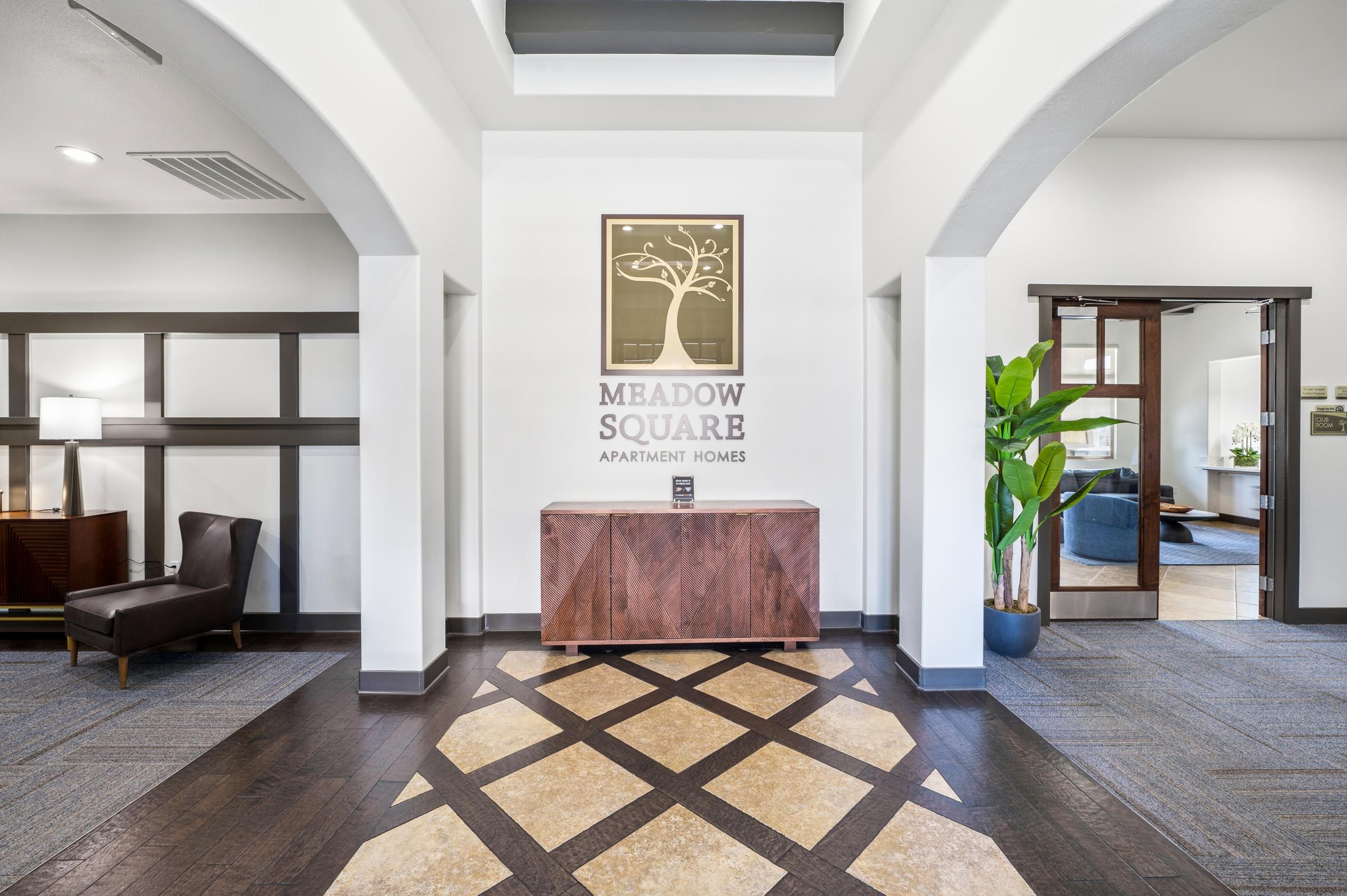 Indoor lobby area of Meadow Square Apartment Homes featuring a decorative sign on the wall, a wooden reception desk, a leather chair, and a potted plant. The flooring has an intricate tile design, and the space is well-lit and inviting.