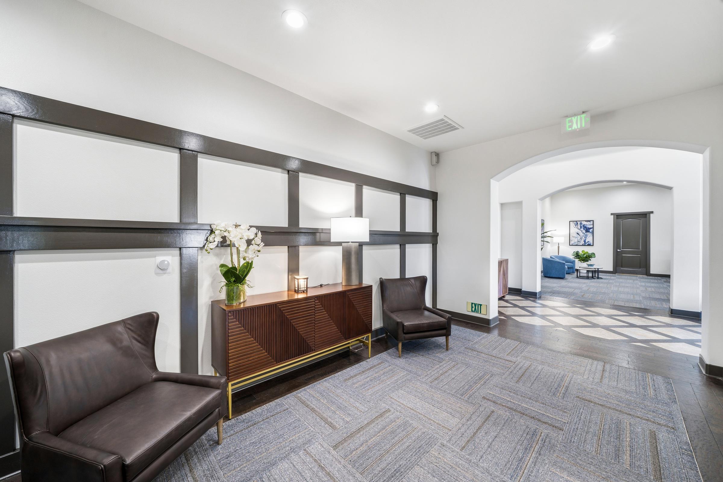 A modern hallway with gray patterned carpet. On the left, two stylish dark chairs are positioned near a sleek wooden console table with a lamp and a potted orchid. The walls feature elegant paneling. An arched doorway leads to another area, visible in the background. The space is well-lit and inviting.