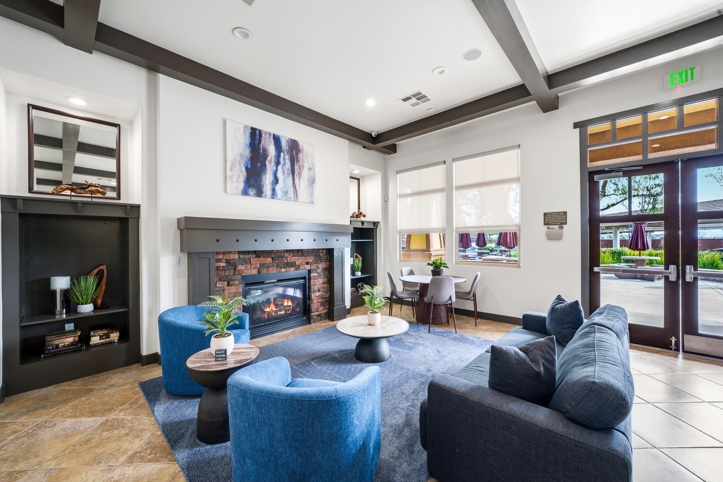 Interior of a modern lounge area featuring a cozy fireplace, blue upholstered furniture including chairs and a sofa, a round coffee table, and large windows letting in natural light. Decor includes a large abstract painting and potted plants. The space is inviting and designed for relaxation and socializing.
