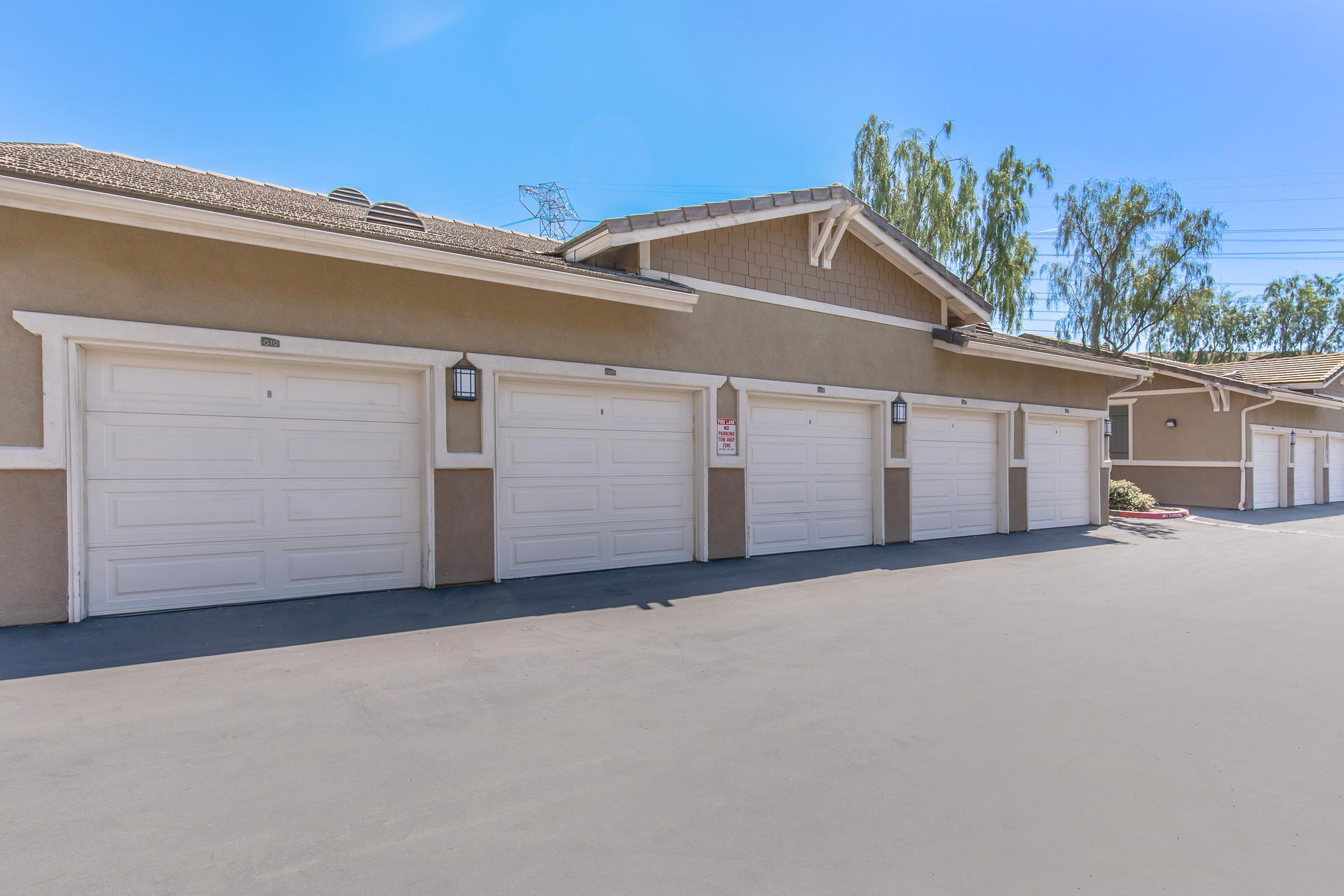 Row of garage doors in a residential area, with a clear blue sky above and landscaping visible in the background. The garages are part of a larger building structure and feature white doors and light fixtures, creating a neat and organized appearance.