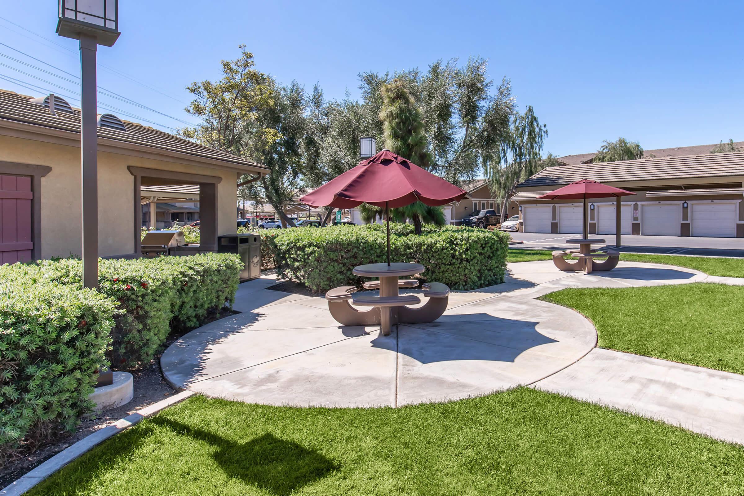 A vibrant outdoor area featuring a well-maintained lawn, concrete pathways, and two picnic tables with maroon umbrellas. Surrounding the space are neatly trimmed bushes and trees, creating a pleasant environment with clear blue skies above.