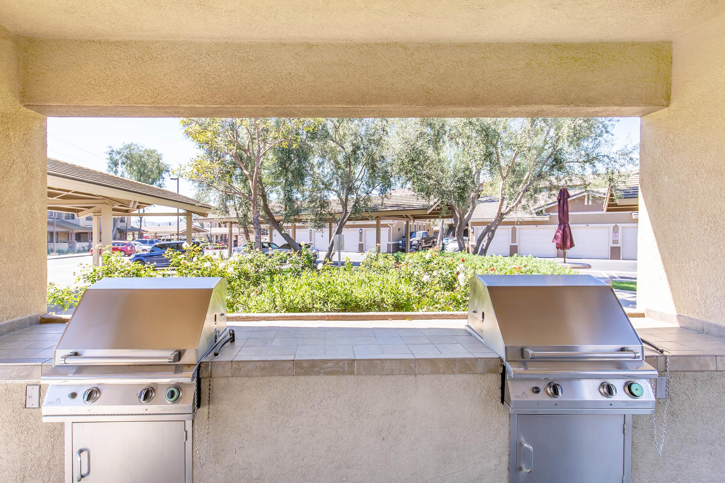 Two outdoor grills positioned side by side under a covered area, looking out onto a landscaped courtyard with trees and greenery. The space is well-lit with natural light and includes a view of a parking area and buildings in the background.