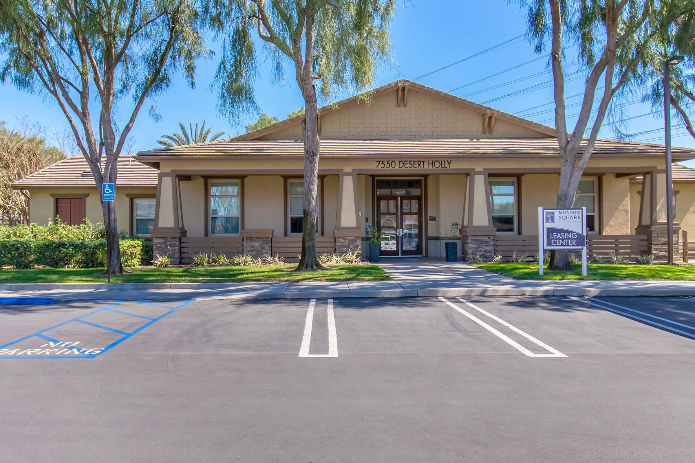 A single-story building with a welcoming entrance featuring large windows and a centered door. The sign in front reads "7550 Desert Holly Learning Center." The surrounding area has neatly trimmed trees and a paved parking lot with designated handicapped spaces. The sky is clear and blue.