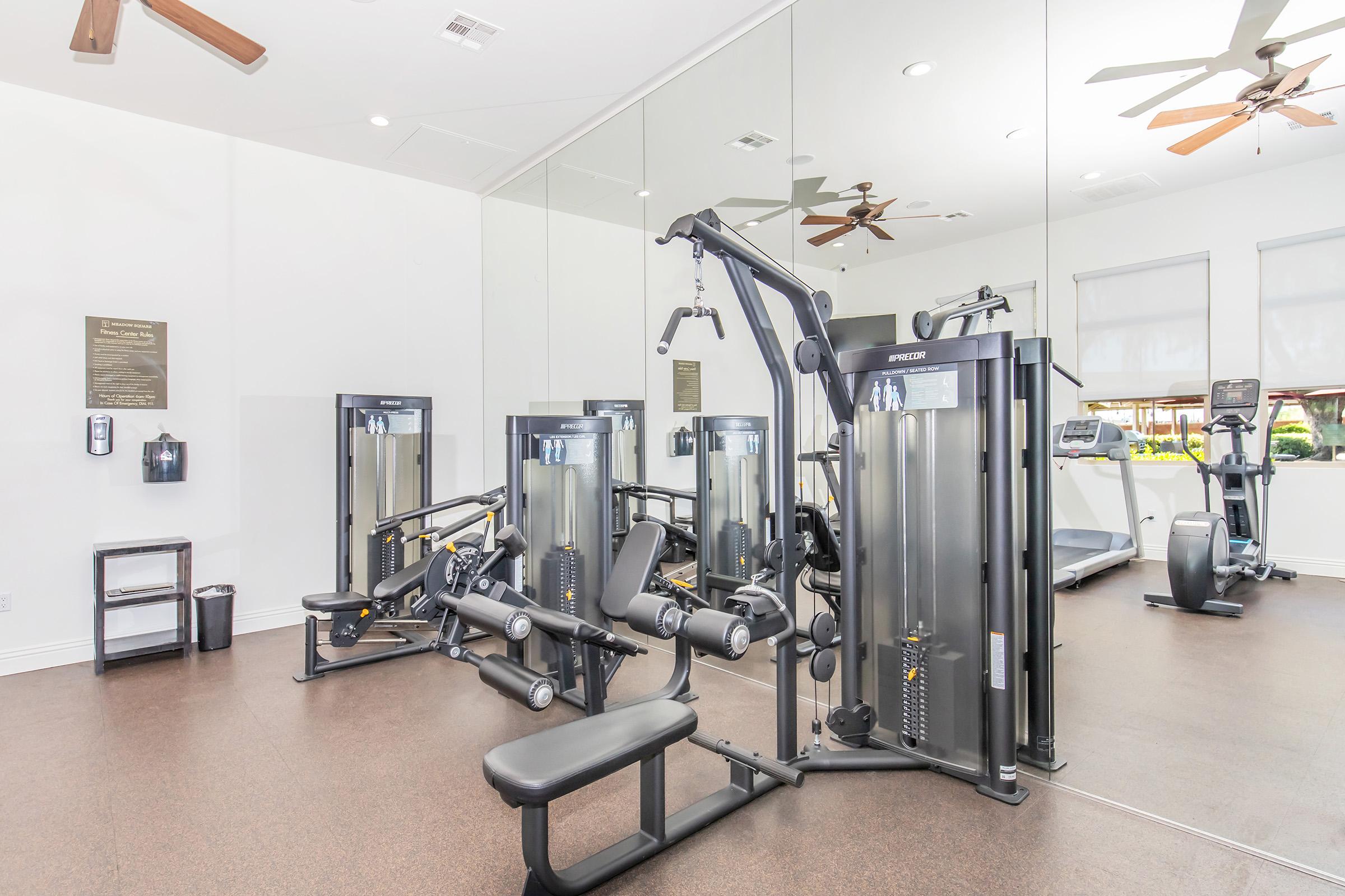 A modern gym interior featuring various workout machines, including weightlifting equipment and cardio machines. The space has large mirrors on the walls, hardwood floors, and ceiling fans, creating a bright and open atmosphere. Fitness equipment is neatly arranged for easy access.