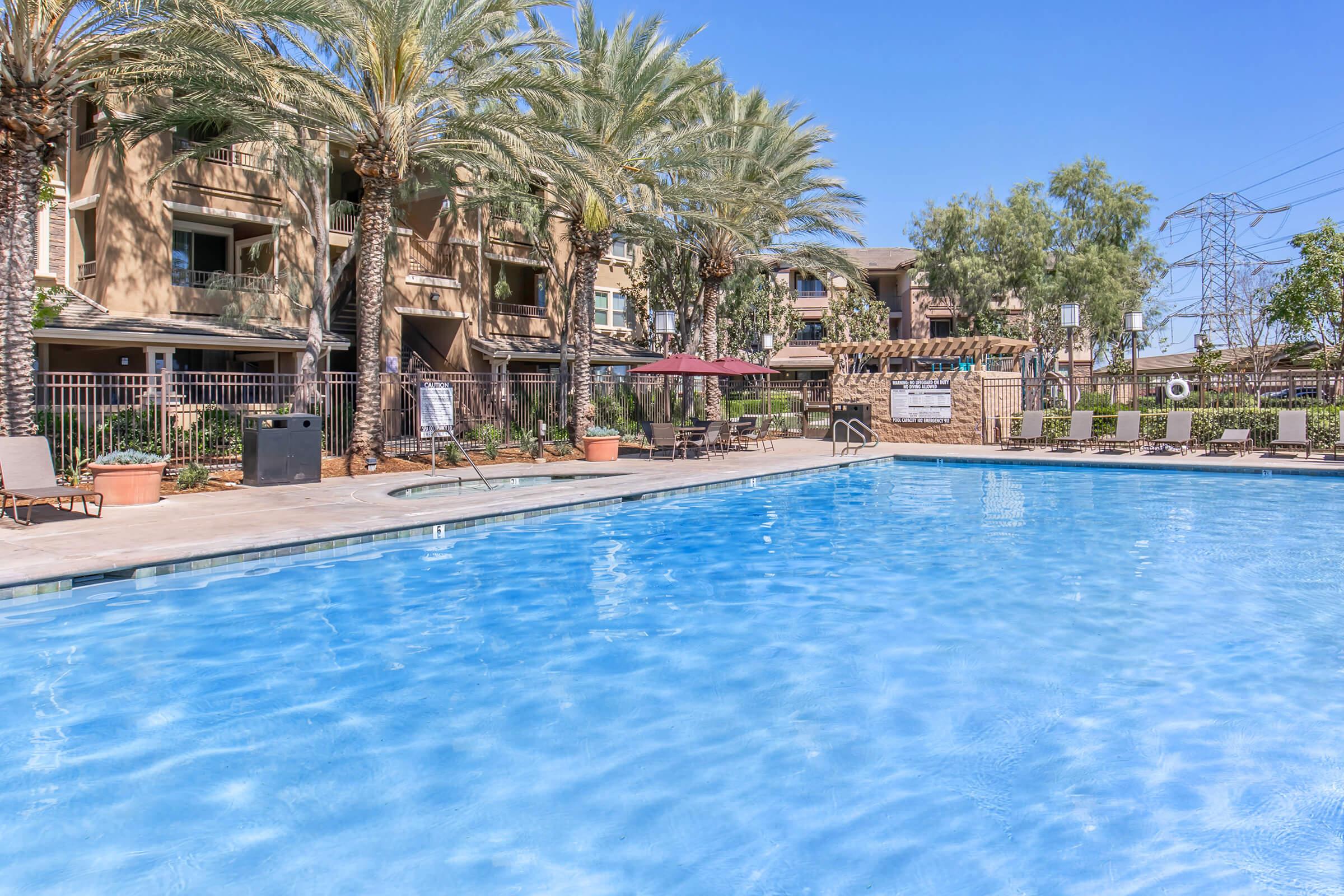 A clear blue swimming pool surrounded by palm trees and lounge chairs. In the background, there are residential buildings and shaded seating areas. The sky is bright and sunny, creating a relaxing outdoor ambiance.