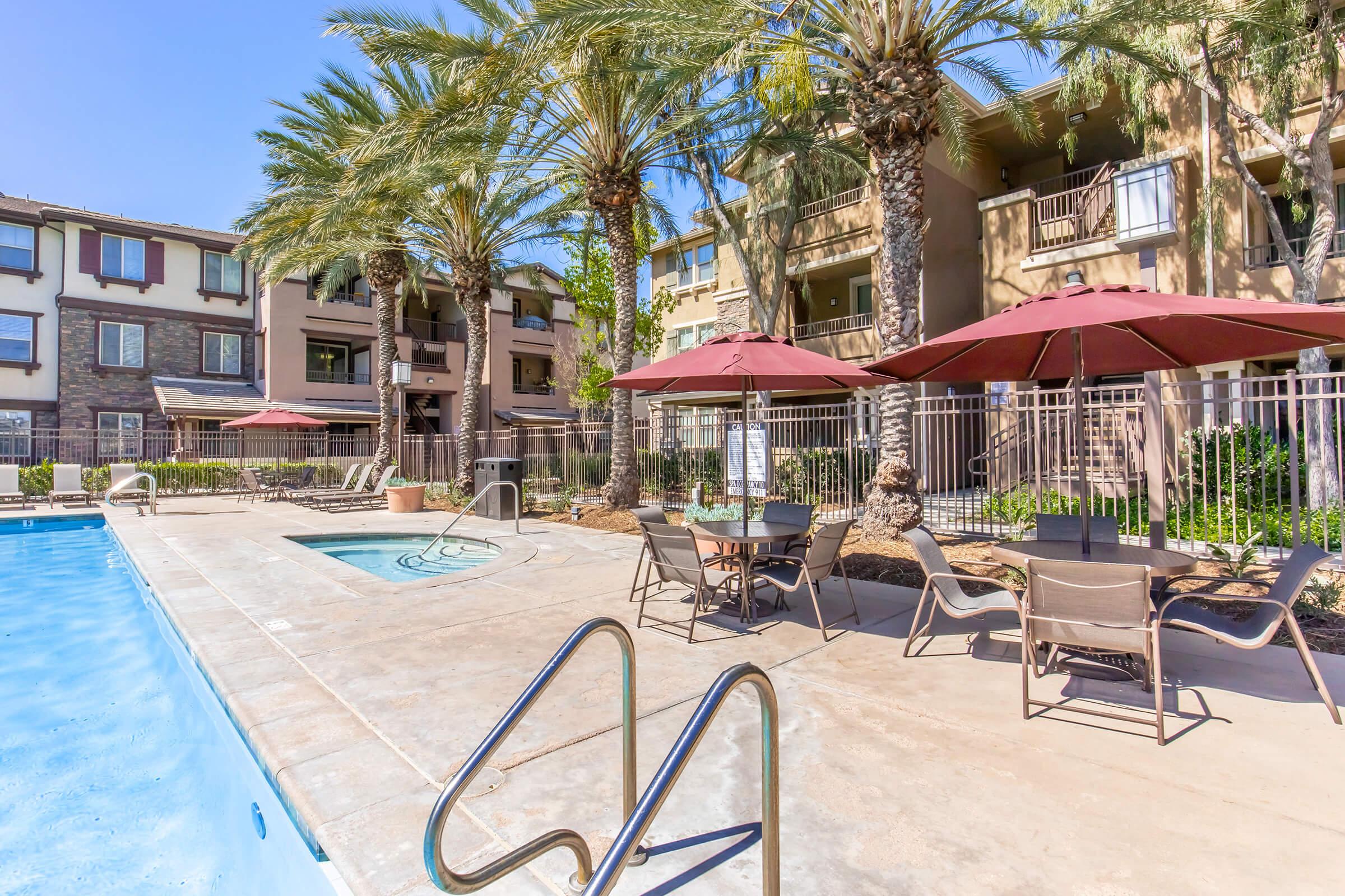 A sunny pool area featuring a clear blue swimming pool, surrounded by palm trees and lounge chairs. There are several umbrellas providing shade over tables, and nearby, a hot tub is situated next to the pool. The backdrop consists of apartment buildings with balconies.