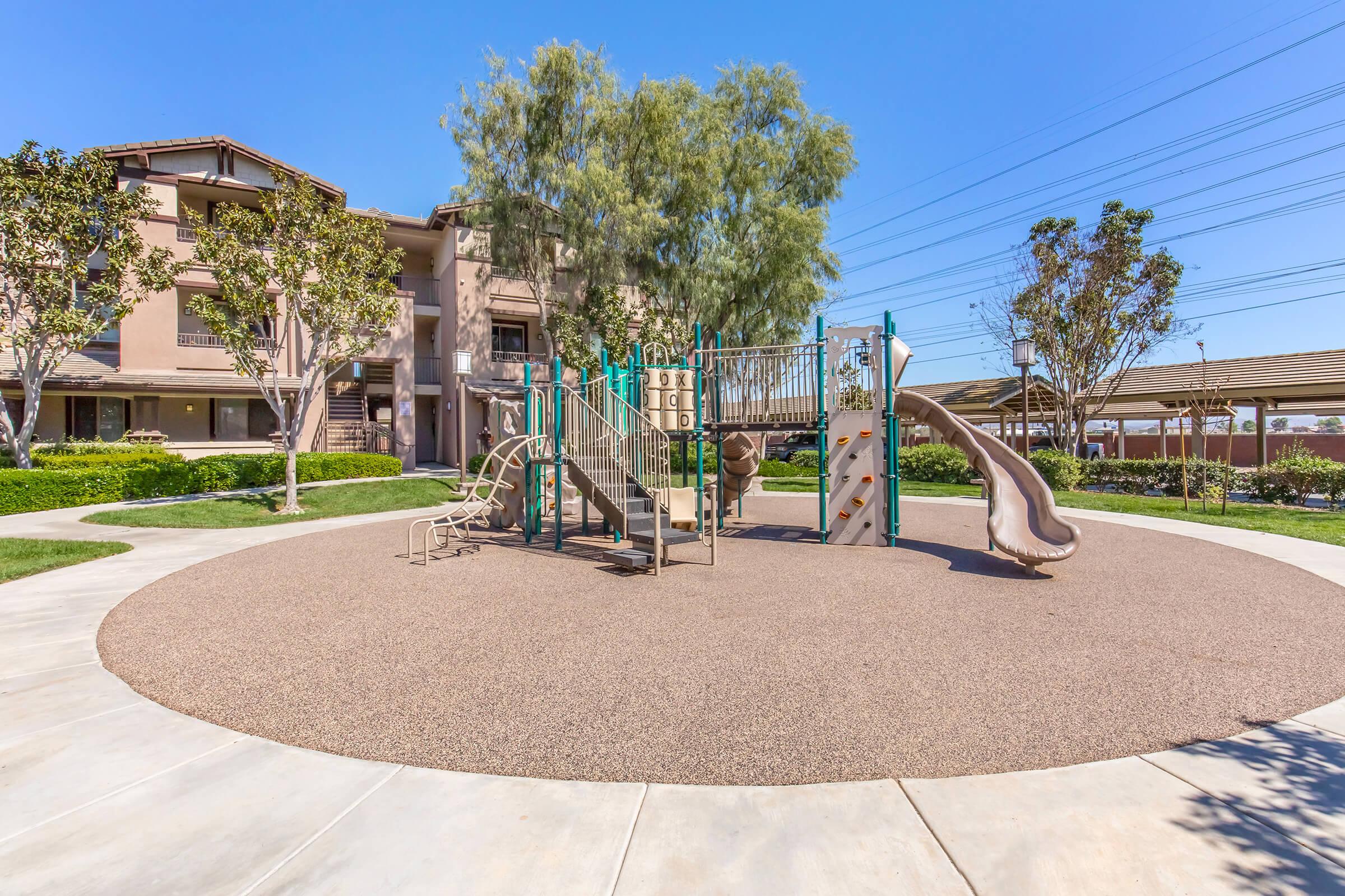 A sunny playground area featuring a colorful play structure with slides and climbing features, surrounded by green grass and trees. Nearby, there are residential buildings and a shaded picnic area, all set against a clear blue sky. Ideal for children's outdoor play.