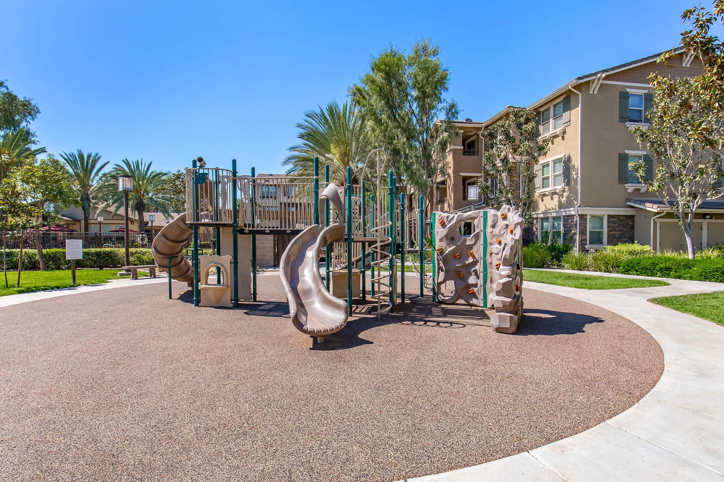 A sunny playground featuring a climbing structure with slides, a rock wall, and various play elements surrounded by green grass and palm trees. Nearby residential buildings are visible in the background, creating a vibrant community atmosphere.