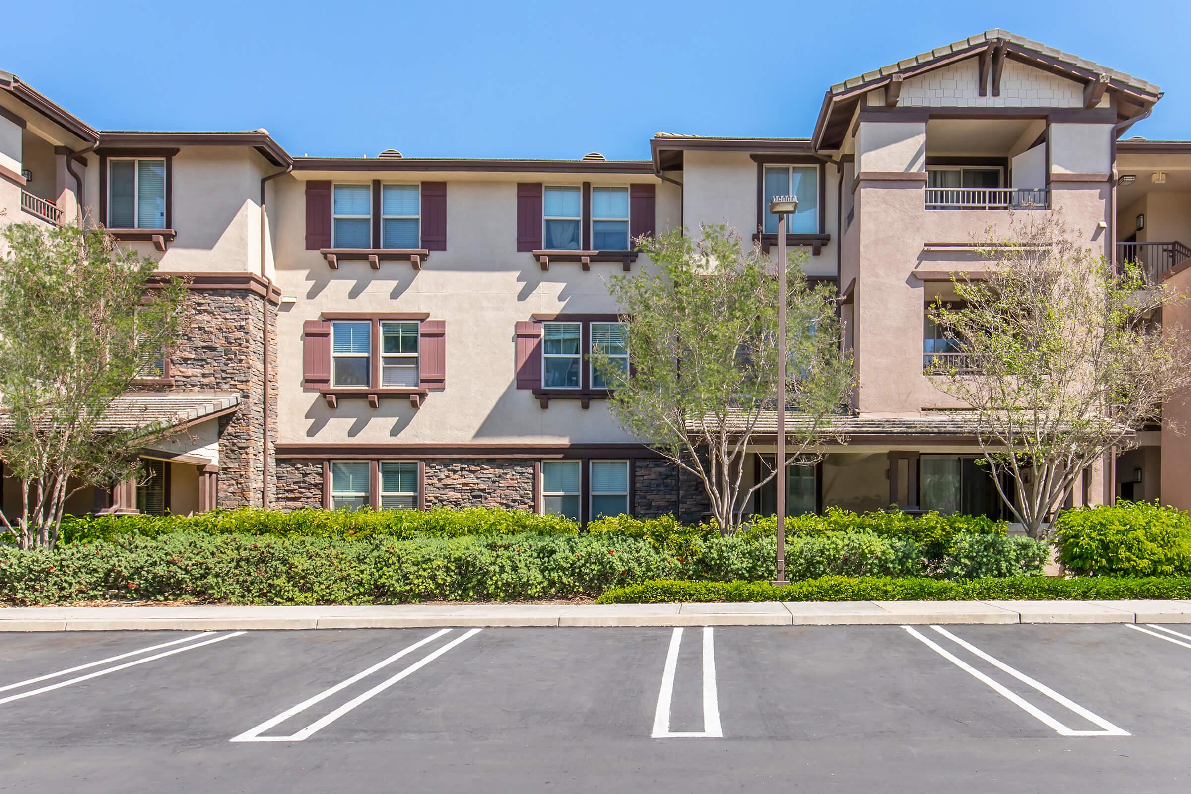 A multi-story residential building featuring a combination of stone and stucco exterior, with several windows and balconies. The front area has neatly arranged parking spaces and landscaping with small trees and shrubs. The sky is clear and bright blue.