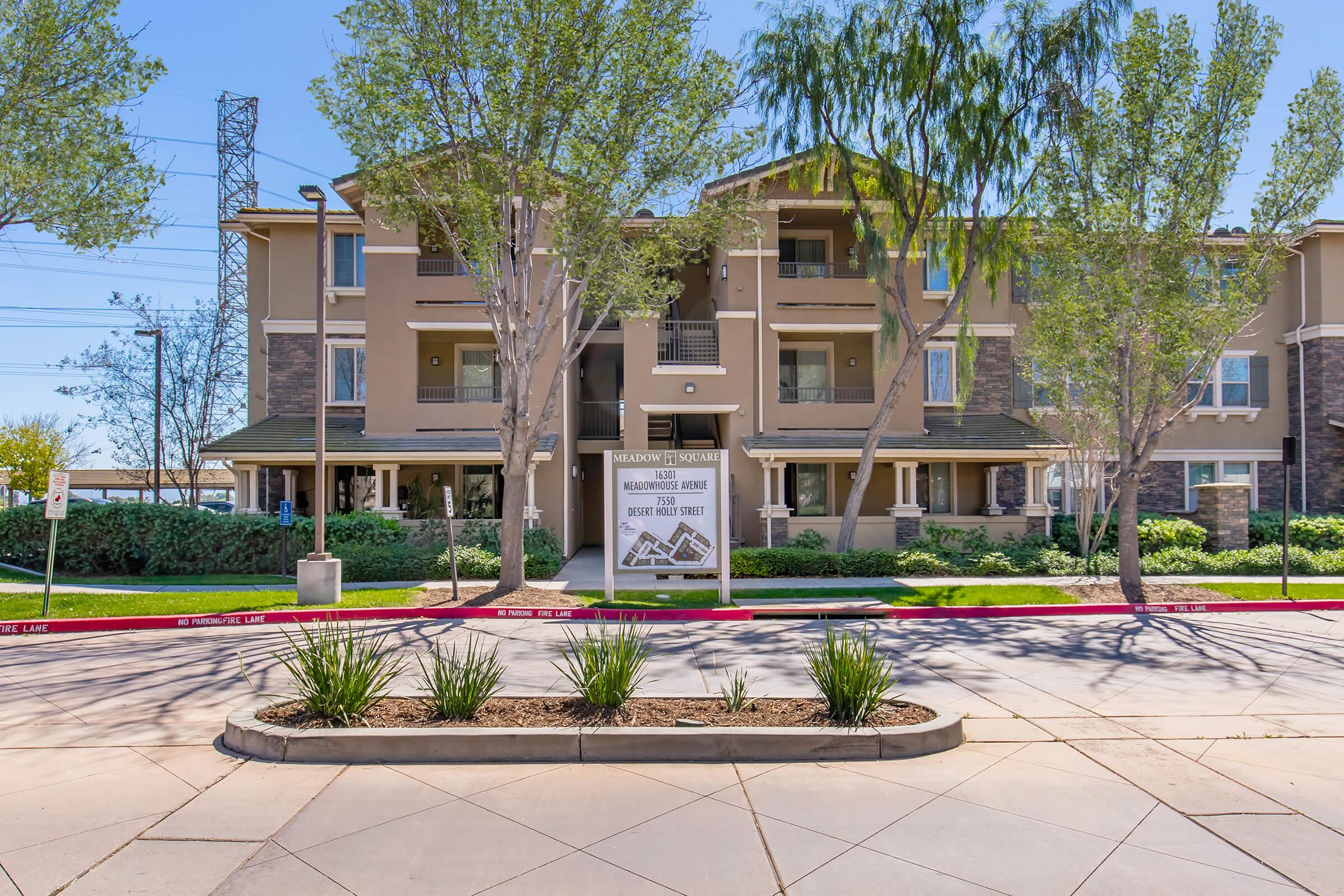 A modern apartment complex with a welcoming sign in front. The building features multiple stories, large windows, and balcony areas. Lush green trees and a neatly maintained landscape enhance the surroundings. The scene is set on a sunny day with a clear blue sky.