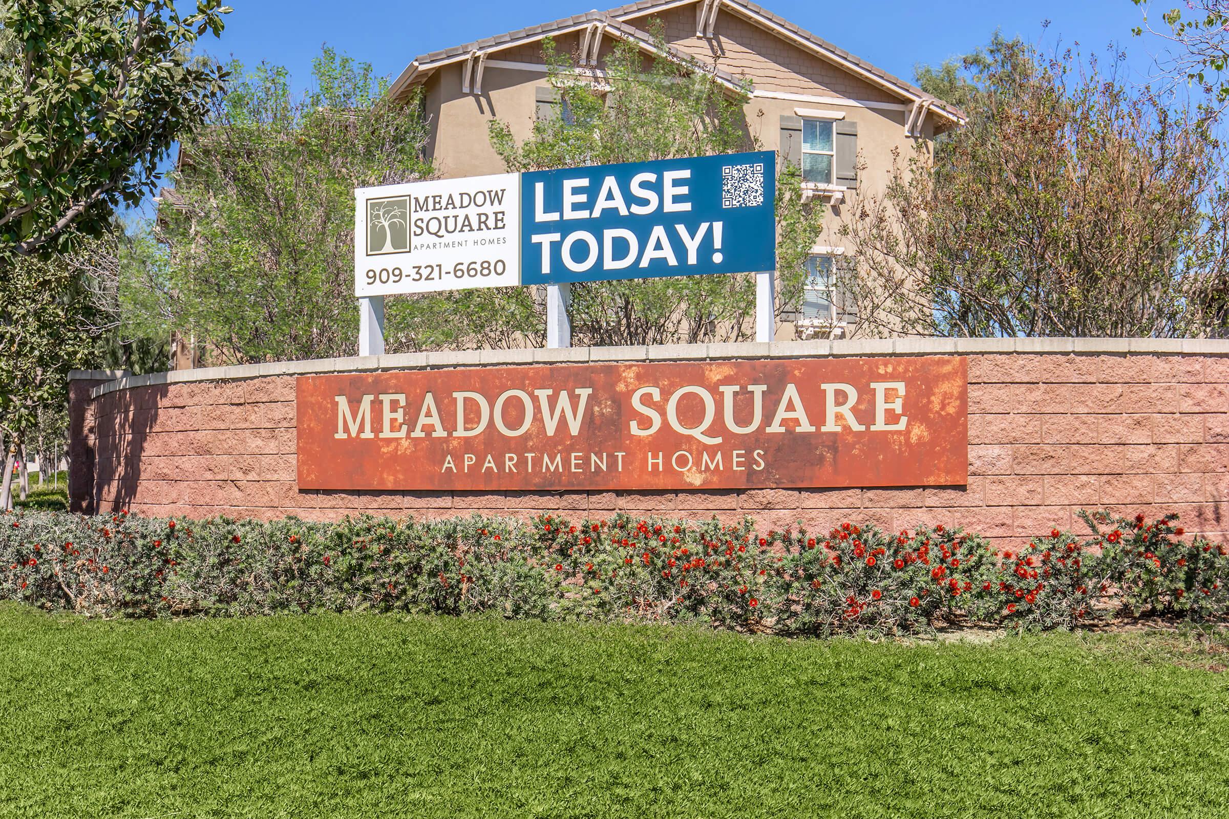 Sign for Meadow Square Apartment Homes featuring the text "LEASE TODAY!" and a contact number. The sign is prominently displayed in front of a landscaped area with greenery and flowers, set against the backdrop of an apartment building. The overall scene conveys a welcoming atmosphere.